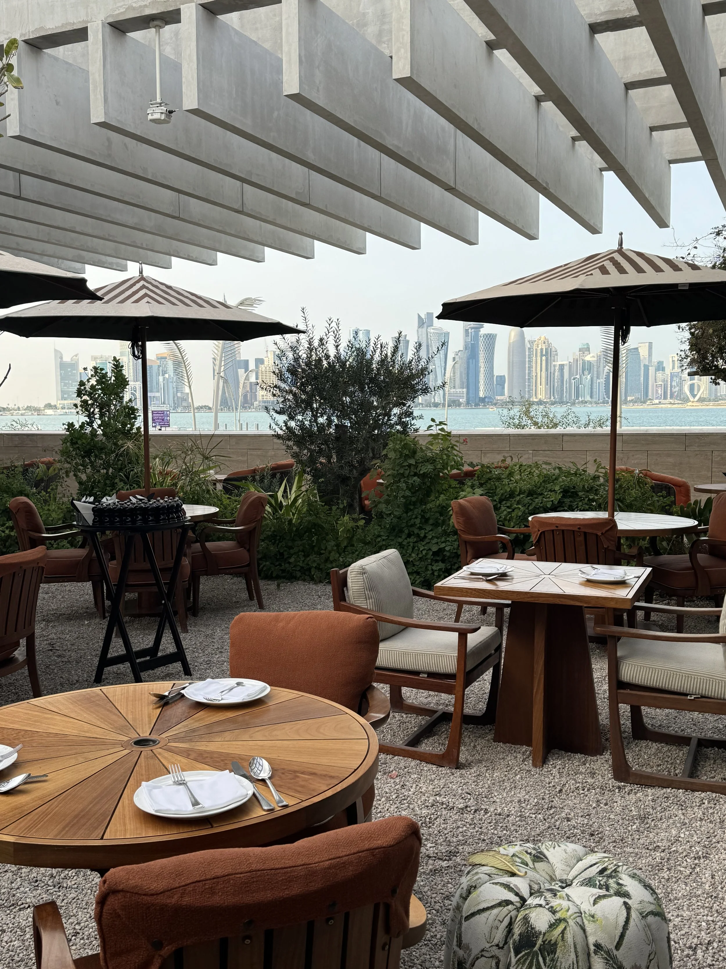 An outdoor restaurant or cafe terrace with wooden tables and cushioned chairs, set with plates and cutlery. There are large umbrellas providing shade, green plants and trees, and a view of a city skyline with tall buildings across a body of water.