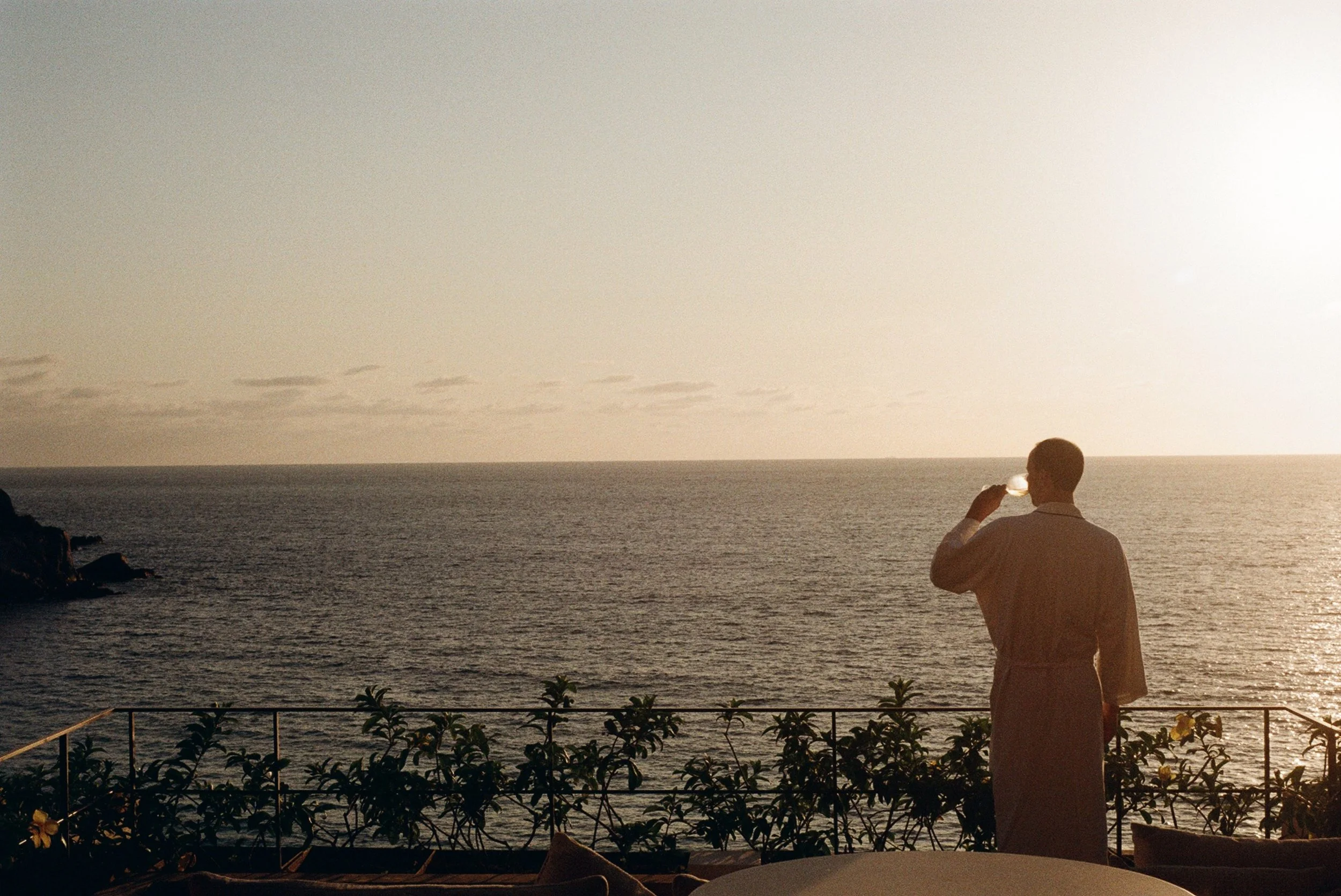 A man in a long-sleeved shirt stands on a balcony holding a drink and enjoying the view of the ocean at sunset.