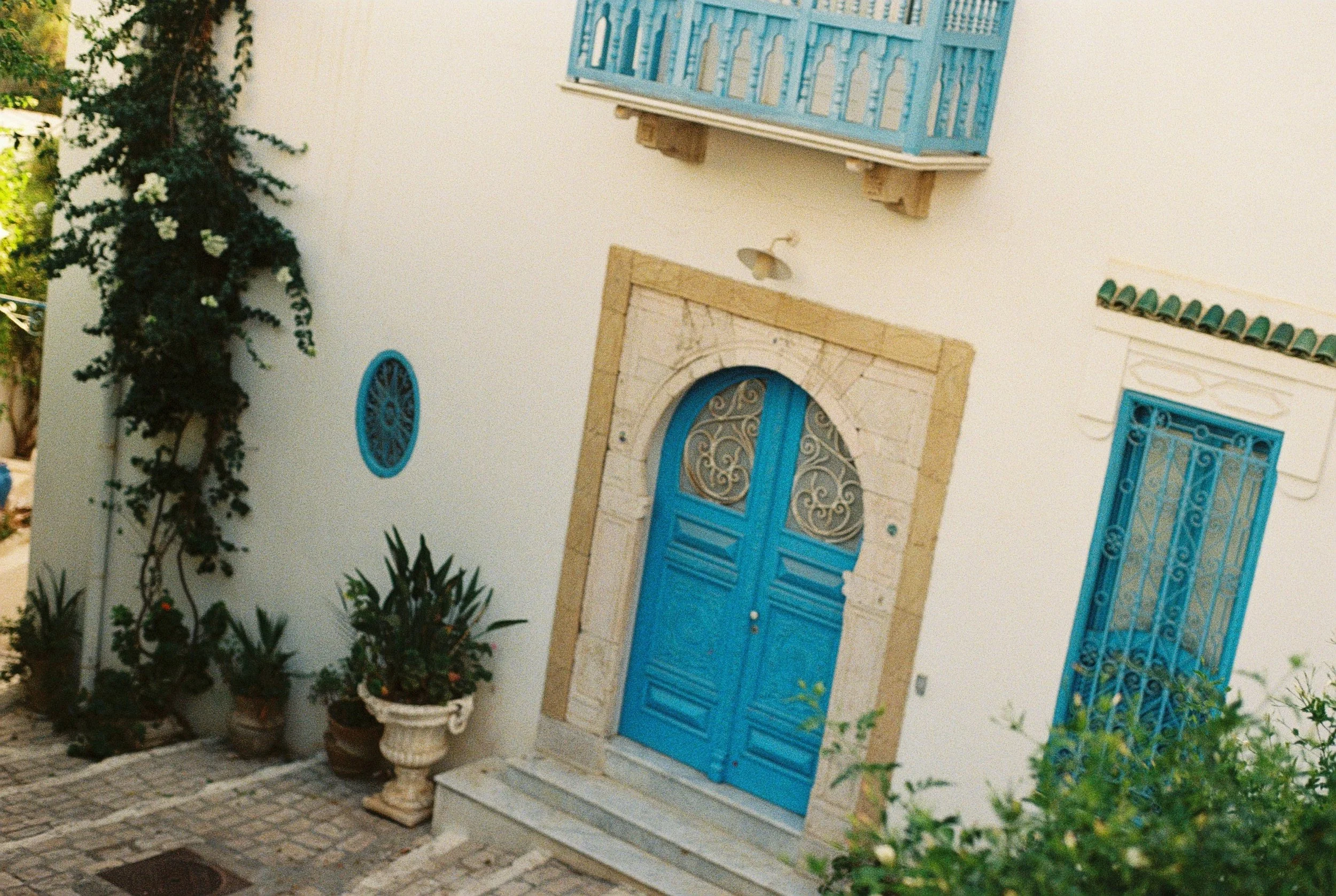 White building with blue doors and windows, surrounded by plants and flowers, cobblestone pathway in front.