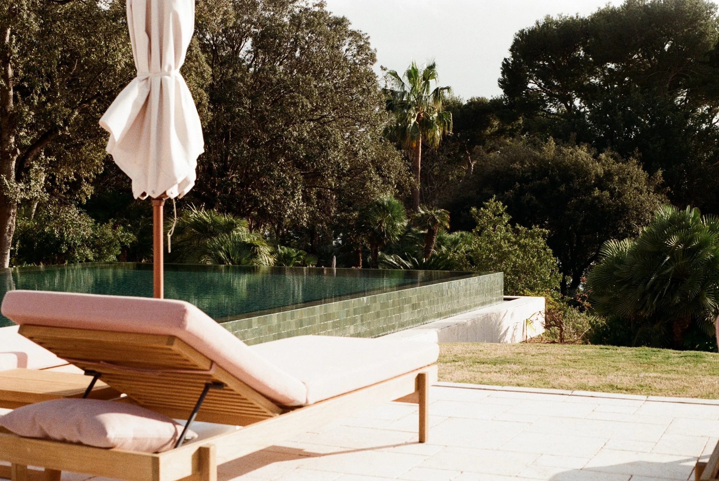 Poolside lounge chairs with cushions and a white umbrella against a backdrop of trees including palm trees.