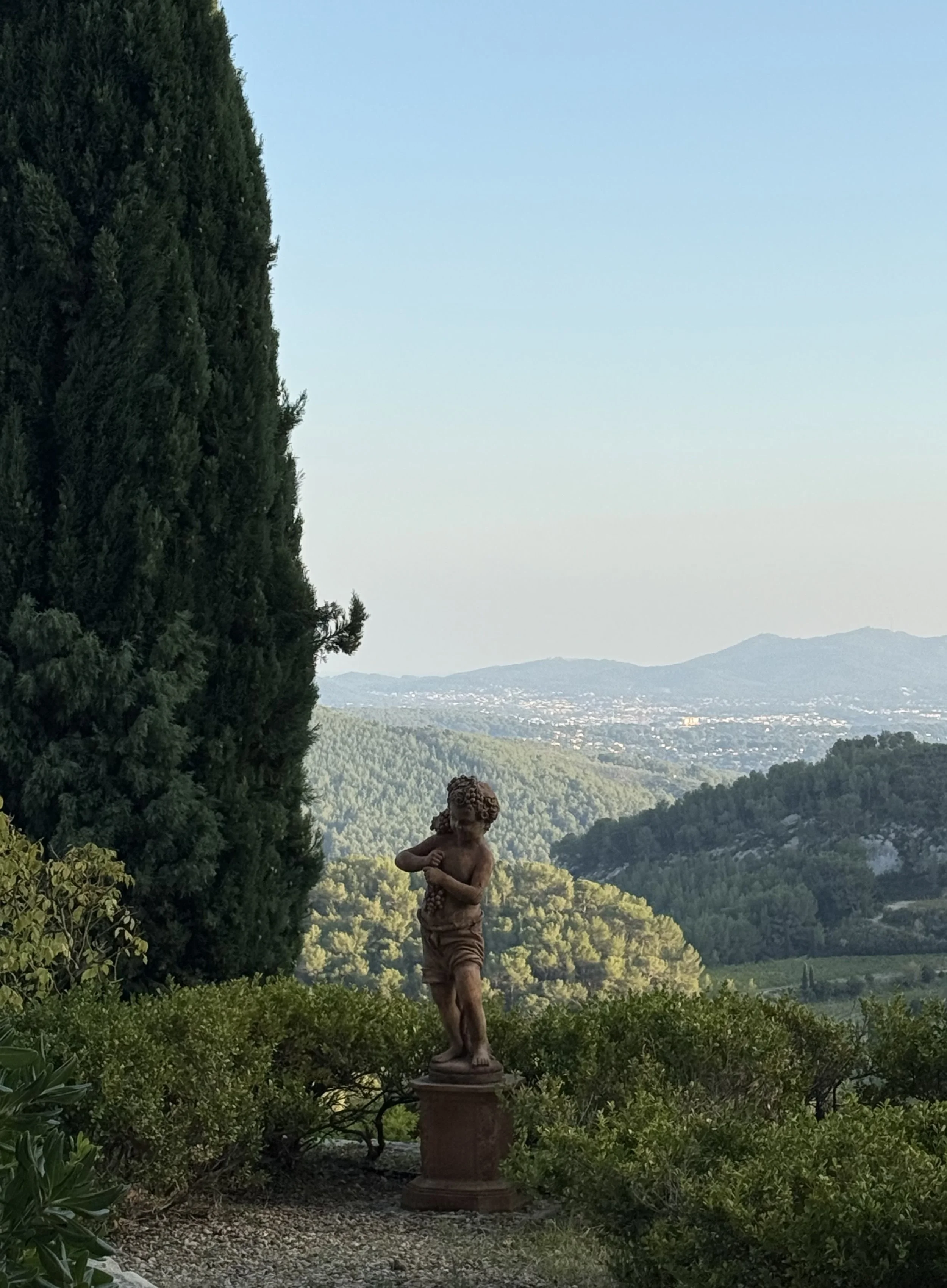 A garden scene with a classical-style statue of a woman holding a small child, surrounded by lush greenery and overlooking distant mountains and a valley.