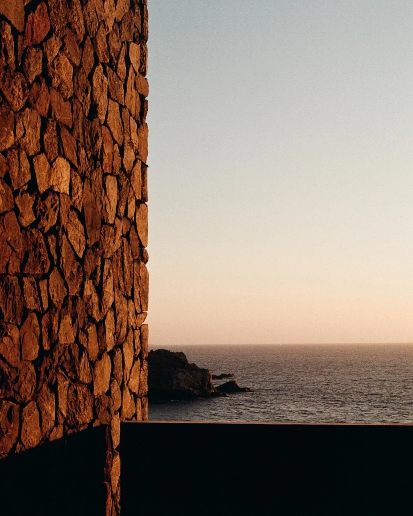 A stone wall is on the left with an ocean view on the right during sunset.