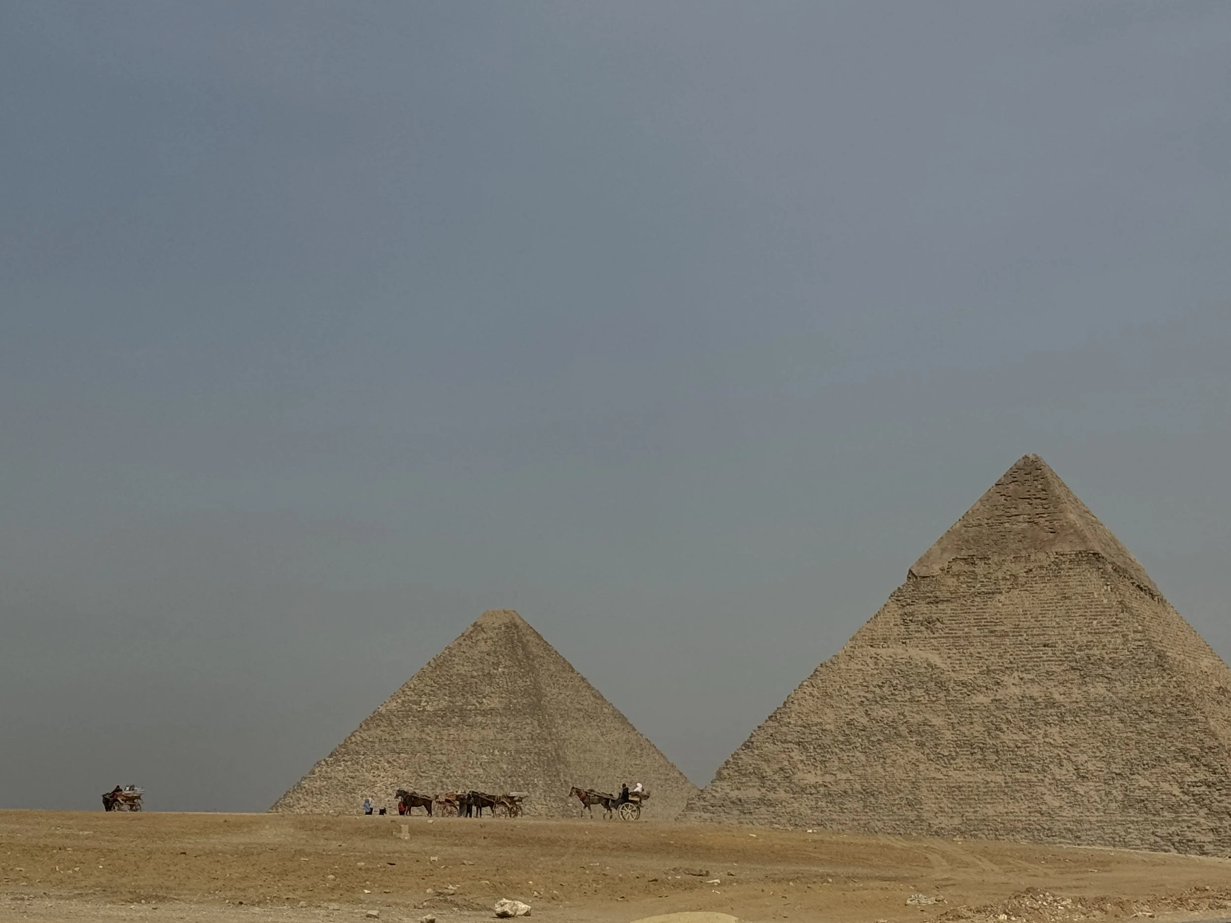 The photo shows two large pyramids in the desert with a group of camels and people in front of them. The sky is clear and blue.
