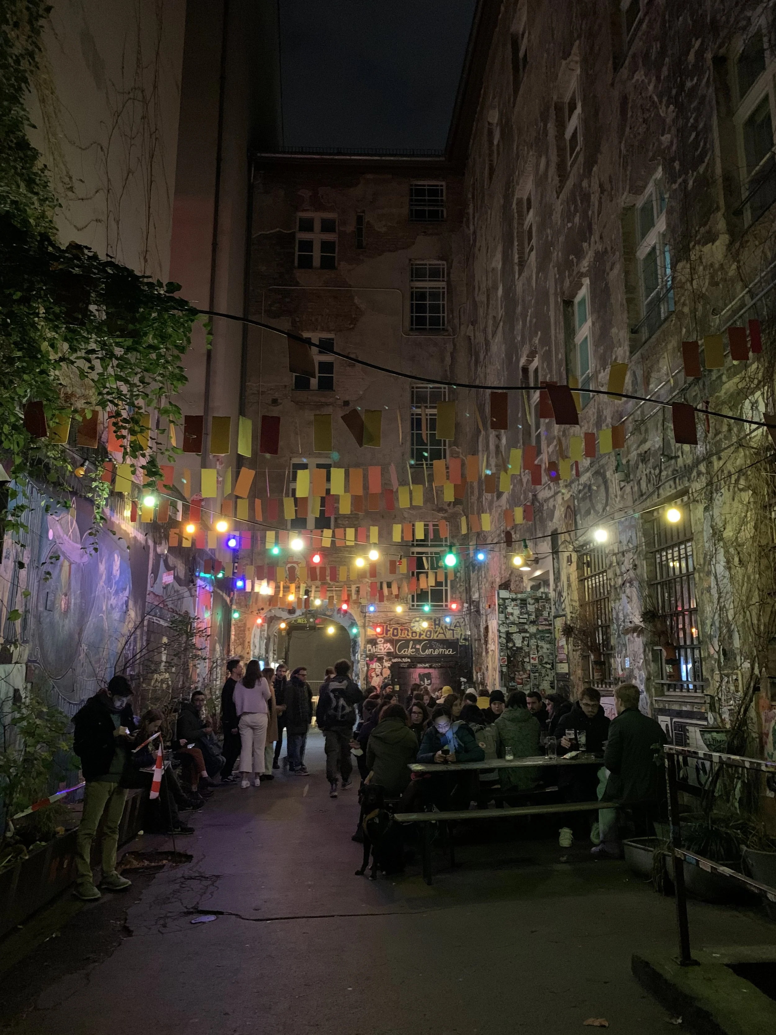 Night scene in a narrow city alley with colorful string lights and hanging paper decorations. People are gathered, sitting at tables and standing, some looking at their phones or chatting. The walls are adorned with street art and posters, creating an artistic and lively atmosphere.