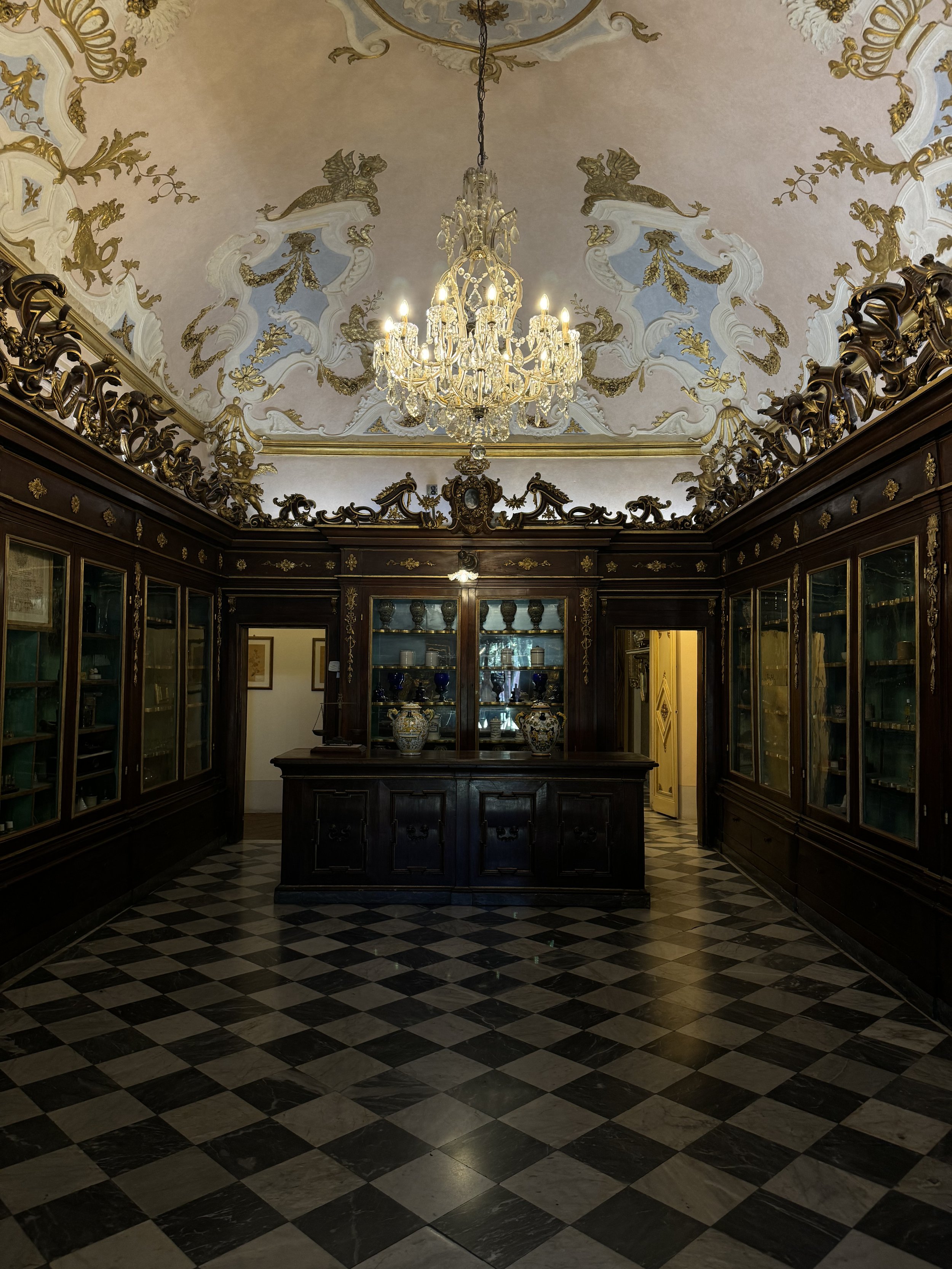 An ornate room with a checkered black and white marble floor, dark wooden built-in cabinets with glass doors, and gold-accented decorations. A large chandelier hangs from a decorative ceiling painted with gold and pale blue accents, featuring mythical creatures and floral patterns.