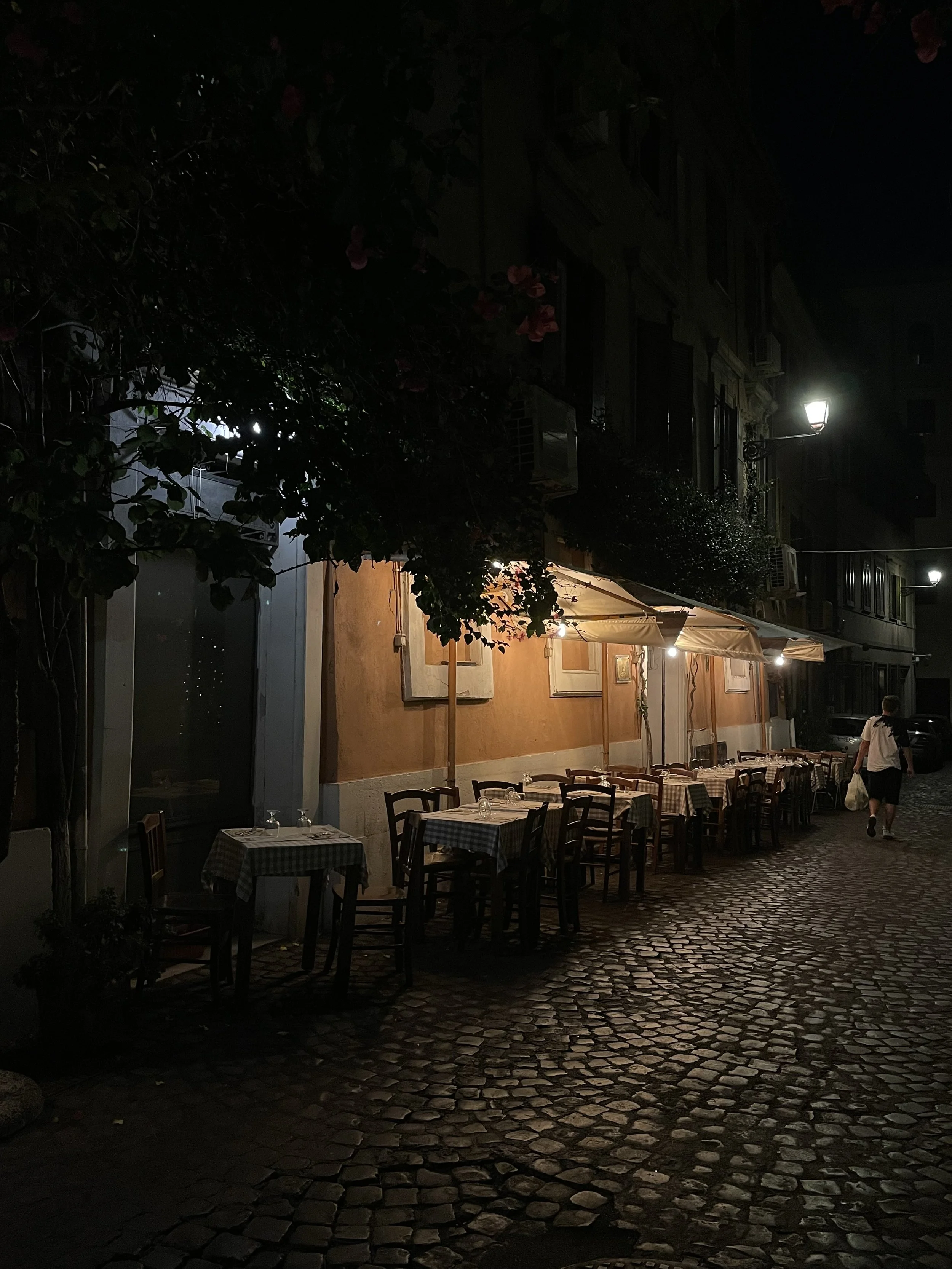 Empty outdoor restaurant with tables and chairs, illuminated by street lamps at night.