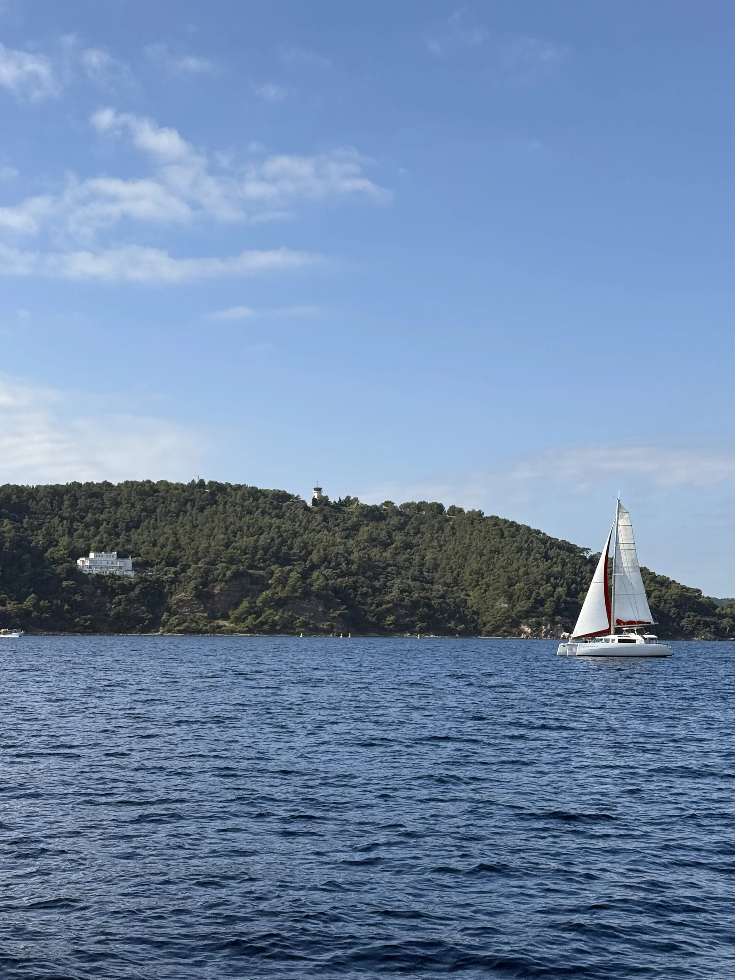 A sailboat with a red and white sail on calm water, with a tree-covered hill and a lighthouse in the background under a partly cloudy sky.