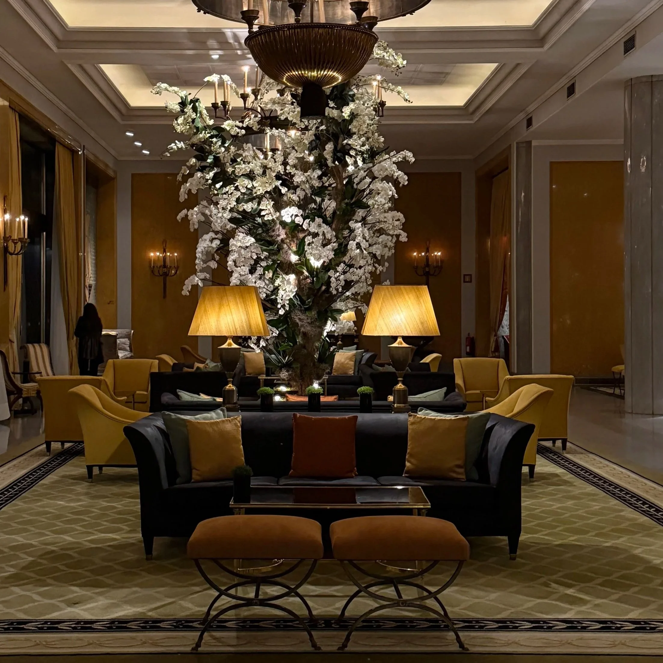 A luxurious hotel lobby with a large floral arrangement of white orchids and dark green leaves in a glass vase, floating on a black table with small decorative plants. The background features yellow couches, gold curtains, and tall wall sconces with lit candles. The ceiling has intricate molding and a grand chandelier.