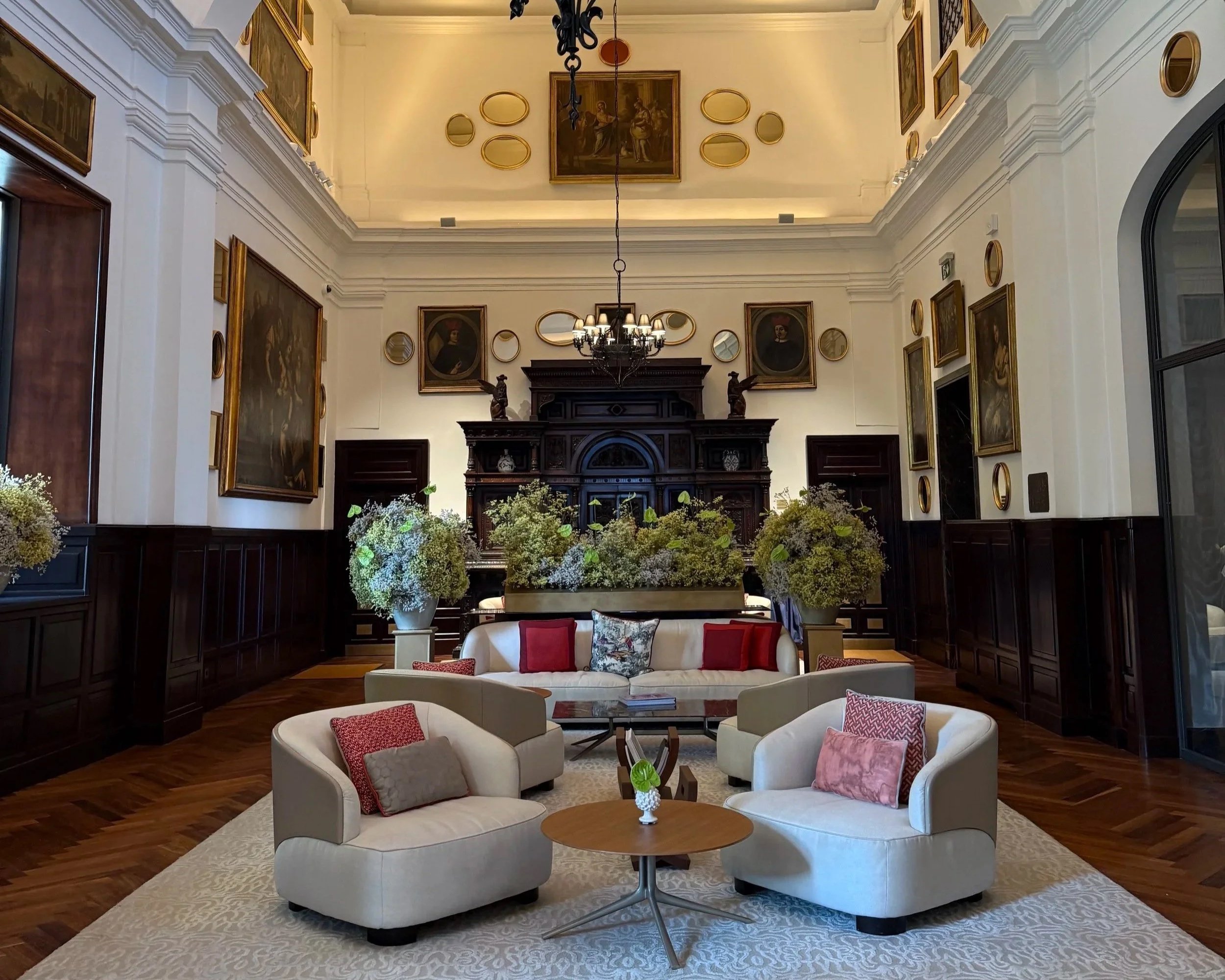 A spacious, elegant living room with high ceilings, adorned with multiple framed paintings and mirrors. The room features a dark wooden fireplace, a chandelier, and various floral arrangements. There are white curved sofas with pink and patterned accent pillows, surrounding a round wooden coffee table on a light patterned rug. Large arched windows allow natural light to illuminate the space.
