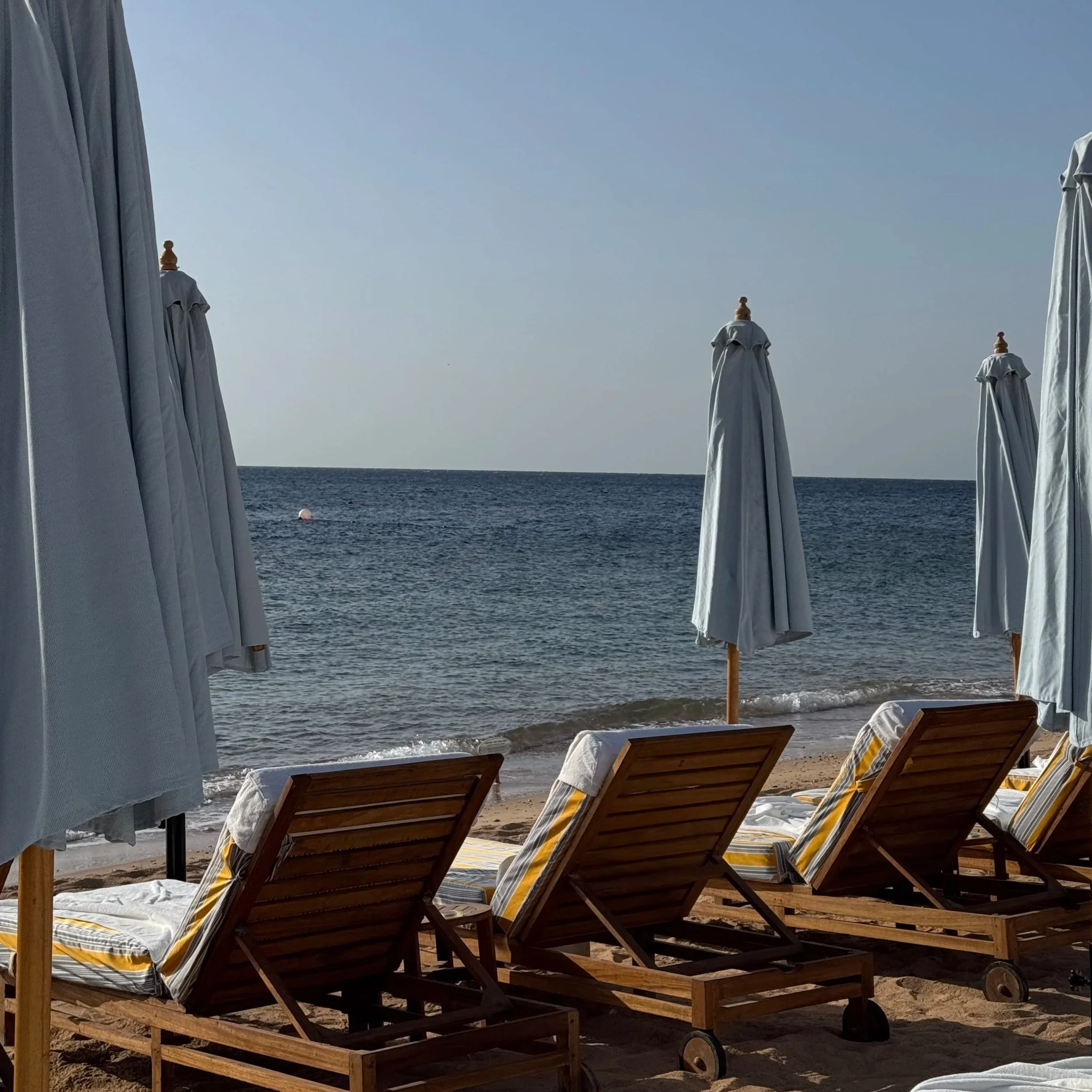 Beach scene seen through open white curtains, with several empty wooden lounge chairs on the sandy beach facing the ocean, and closed white umbrellas in the background under a clear blue sky.