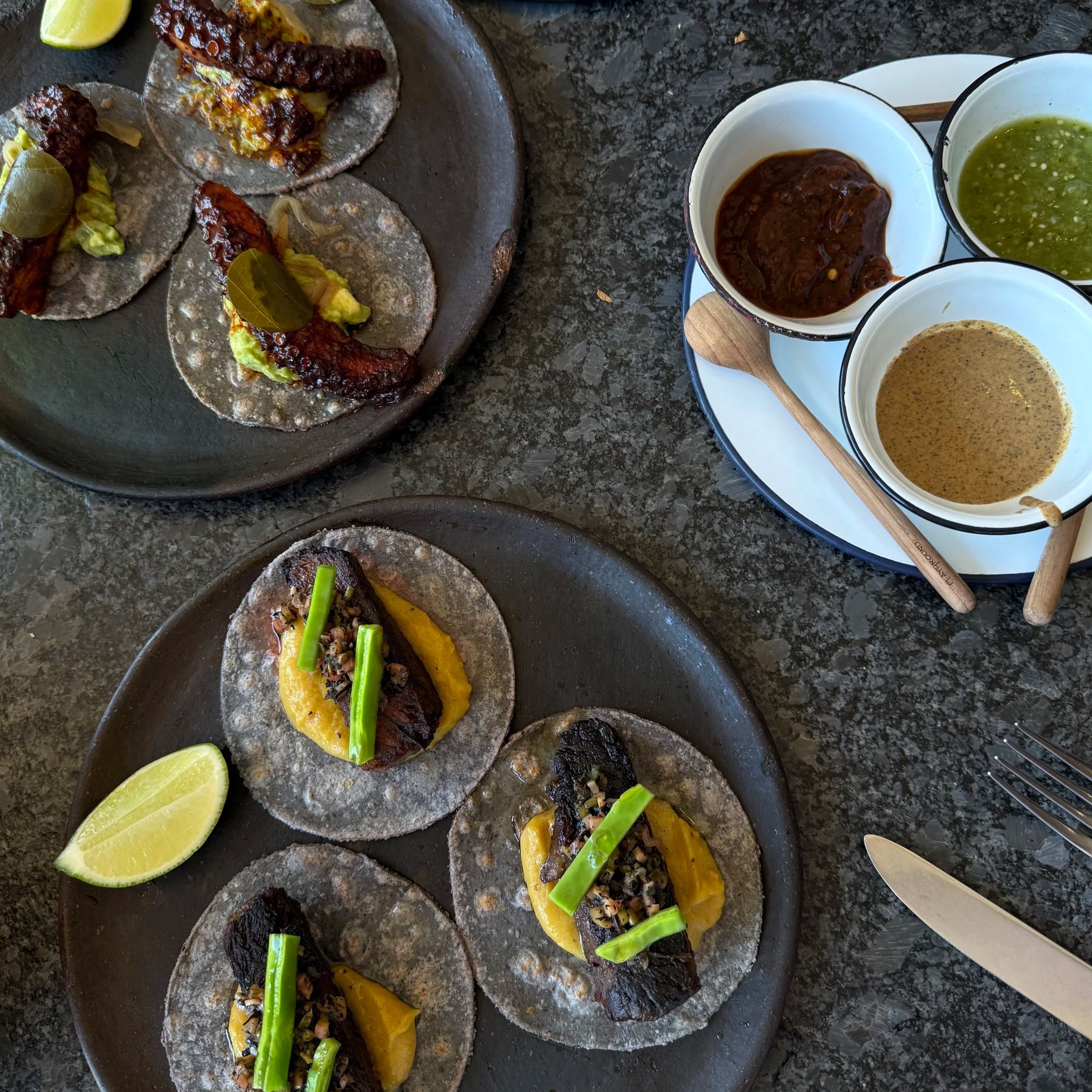 Tacos with grilled meat, topped with green vegetables, served with lime wedges and various sauces on the side.
