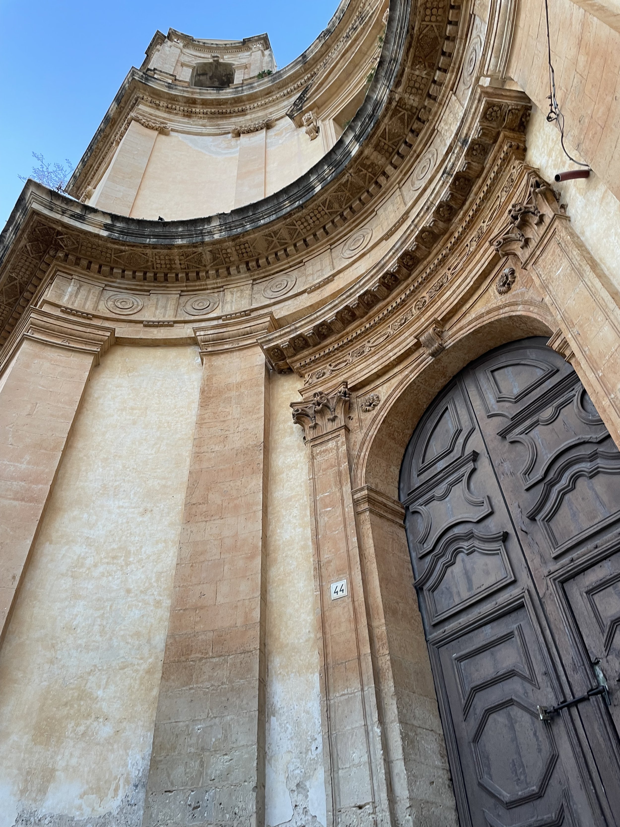 Low-angle view of a historic building's ornate stone facade, part of a curved wall with intricate decorative details, and a large dark wooden door with carved geometric patterns. The blue sky is visible in the background.