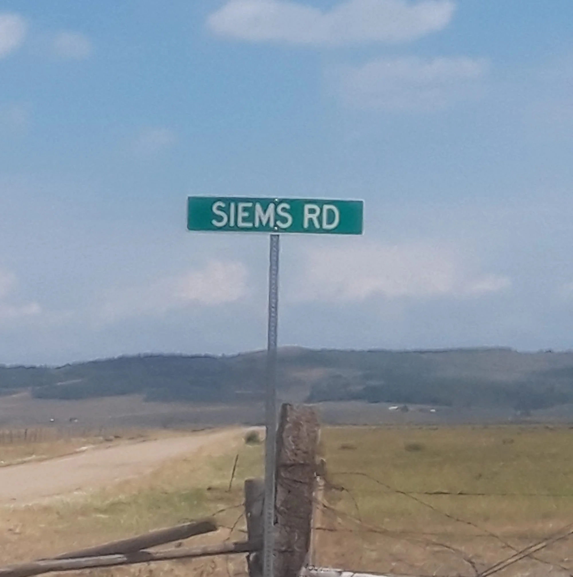 Street sign reading 'SIEMS RD' against a partly cloudy sky, with a utility pole and some grass and dirt in the foreground.