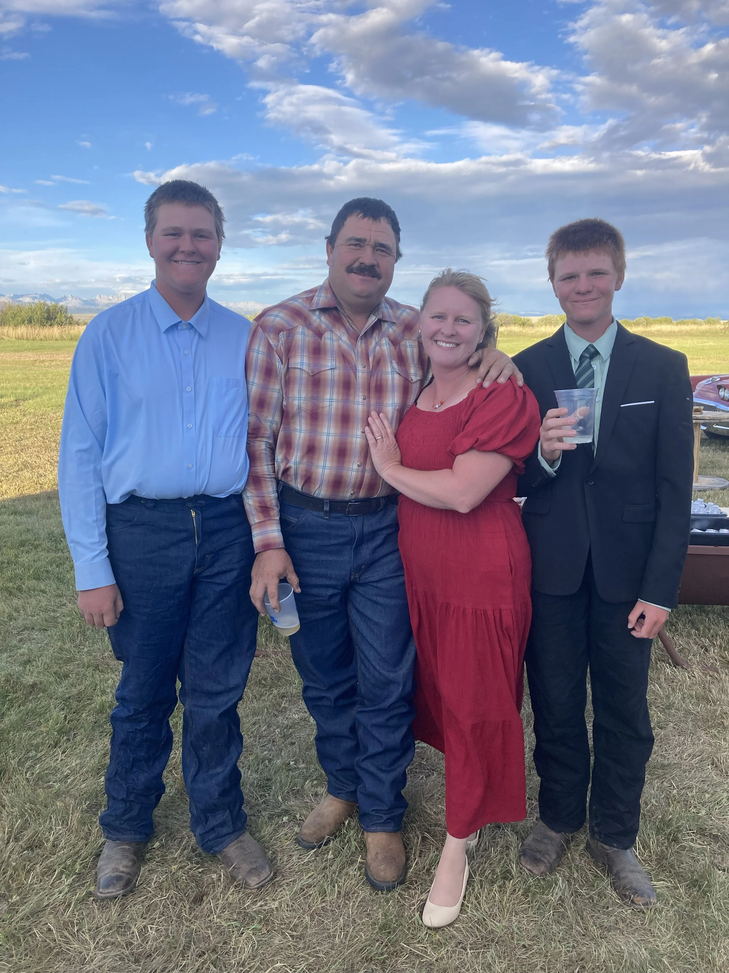 A family of four standing outdoors on a grassy field, smiling. The father has a mustache and is wearing a plaid shirt, the mother has blonde hair and is wearing a red dress, and the two young men are wearing formal attire with jackets and ties. The background shows a partly cloudy sky and distant mountains.
