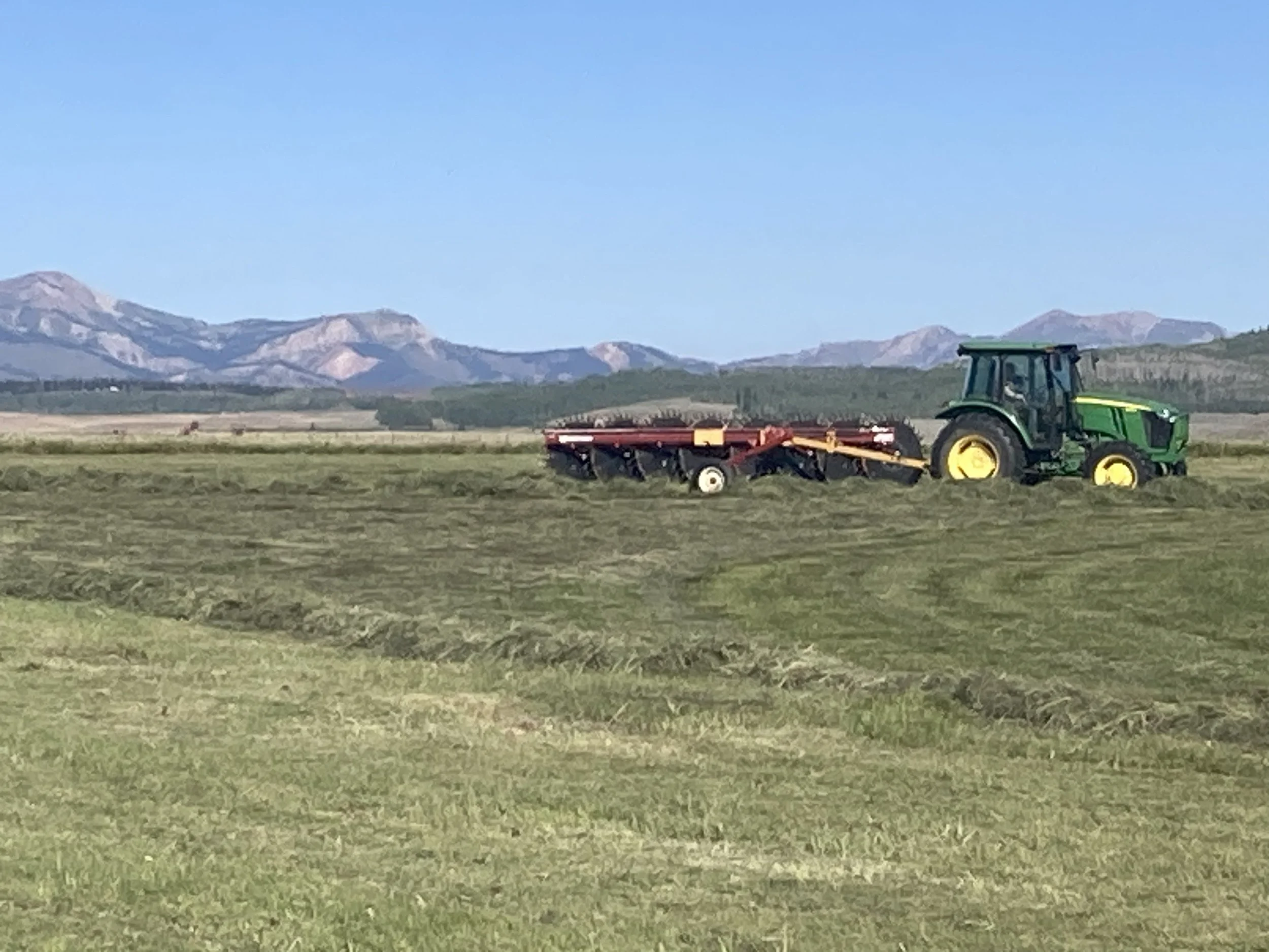 A green tractor pulling a red and yellow plow is working in a field with mountains in the background under a clear blue sky.