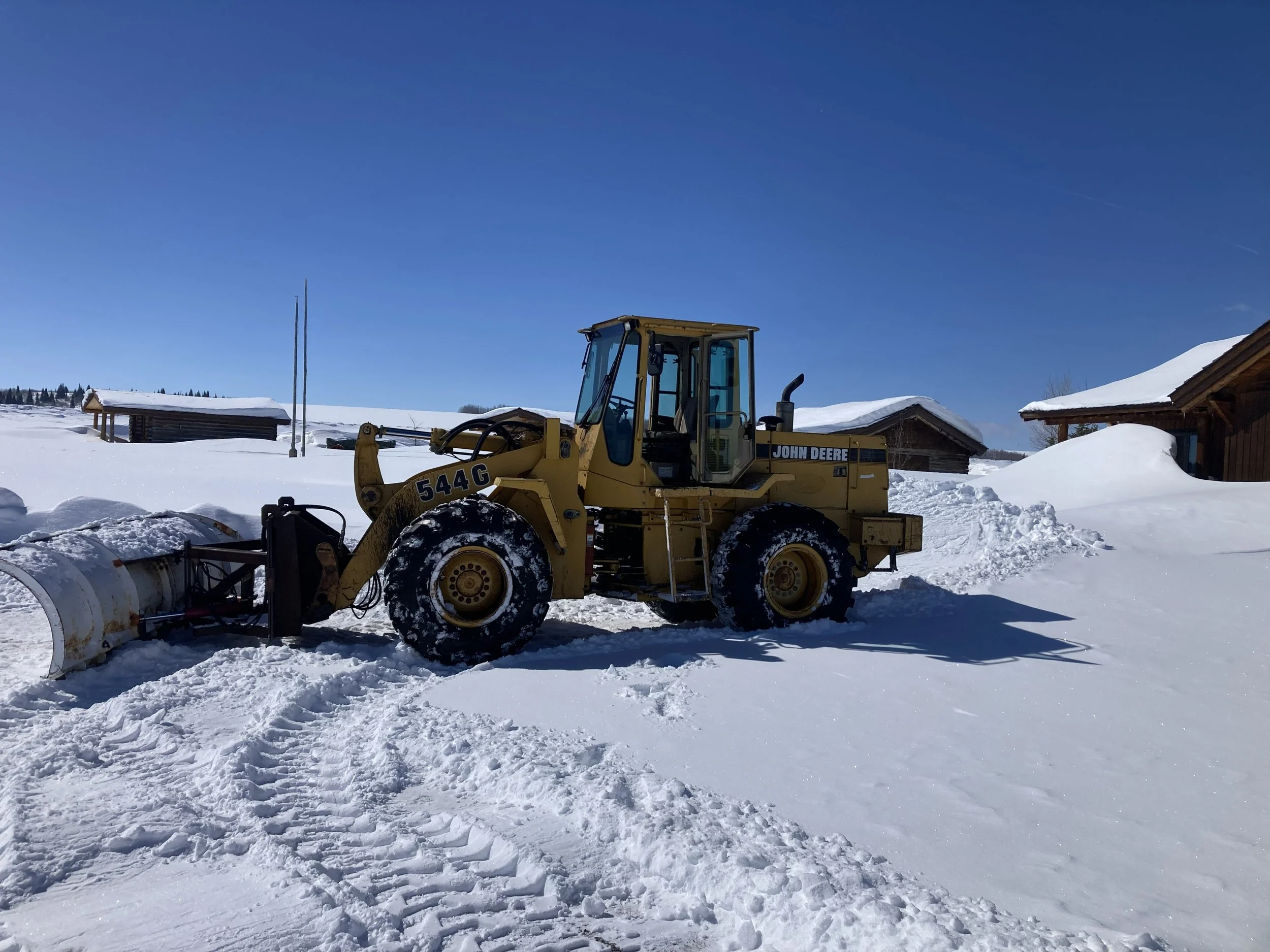 A yellow John Deere snow plow tractor parked in a snowy landscape with small wooden buildings and a clear blue sky in the background.