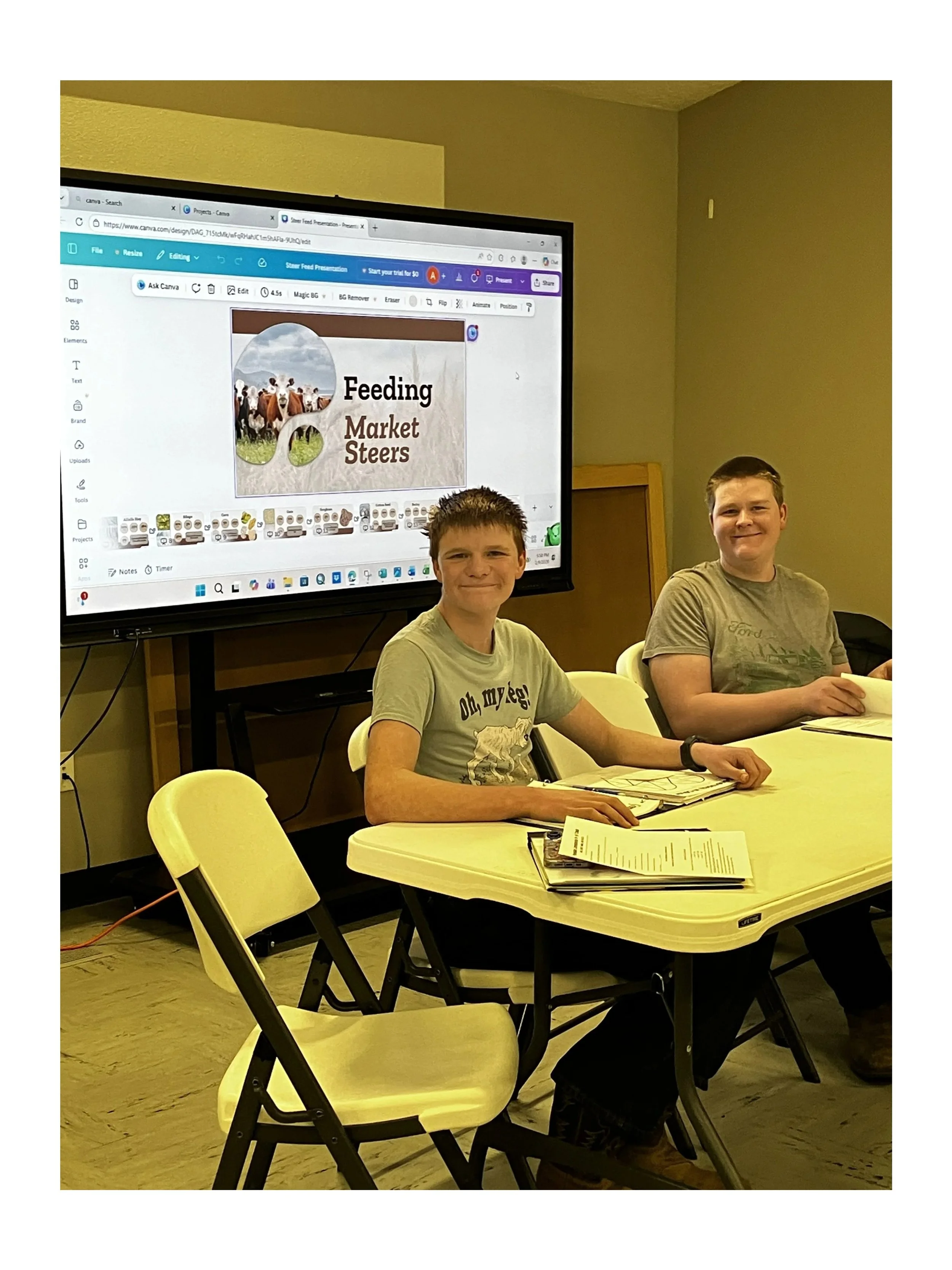 Two young men sitting at a table in a classroom or meeting room with a large screen behind them displaying a presentation slide titled 'Feeding Market Steers' with text and images of cows and a farm scene.