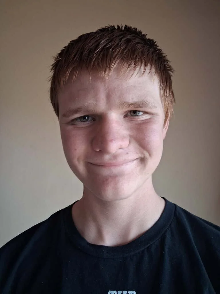 A young boy with red hair and blue eyes smiling at the camera, wearing a black T-shirt, standing against a plain beige wall.