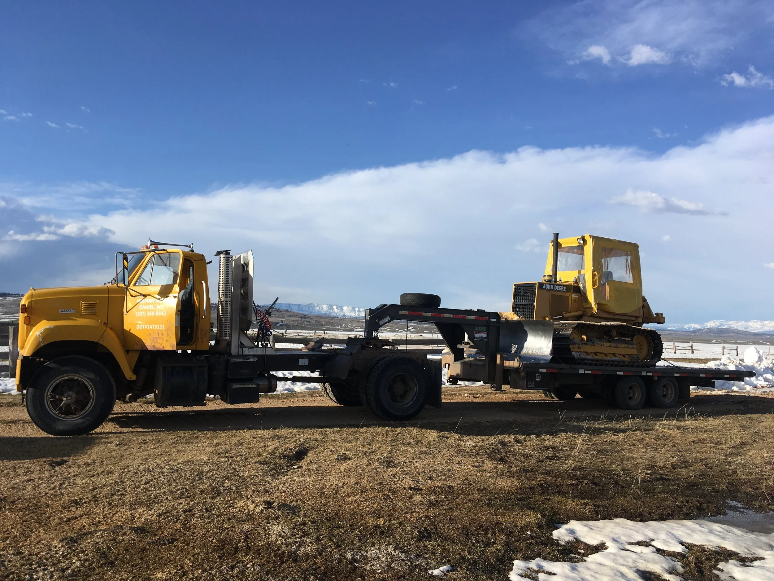 Yellow construction vehicles parked on a flat area with snow, mountains and blue sky in the background.