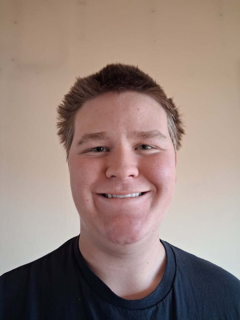 A young man with light skin and short, spiky light brown hair smiling at the camera, wearing a black t-shirt, against a plain beige wall.