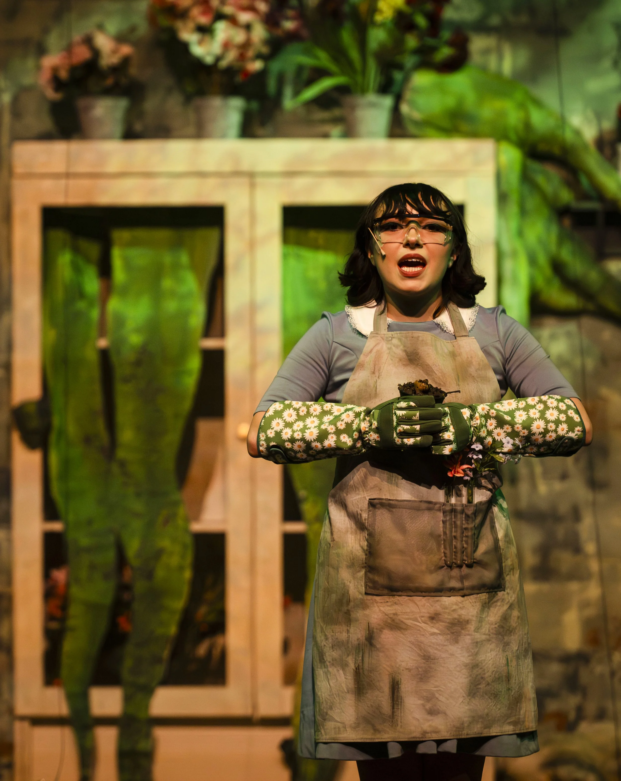 A woman wearing safety glasses and a floral apron stands behind a wooden table holding gardening gloves and a plant. There are potted plants and a wooden structure with hanging plants in the background.