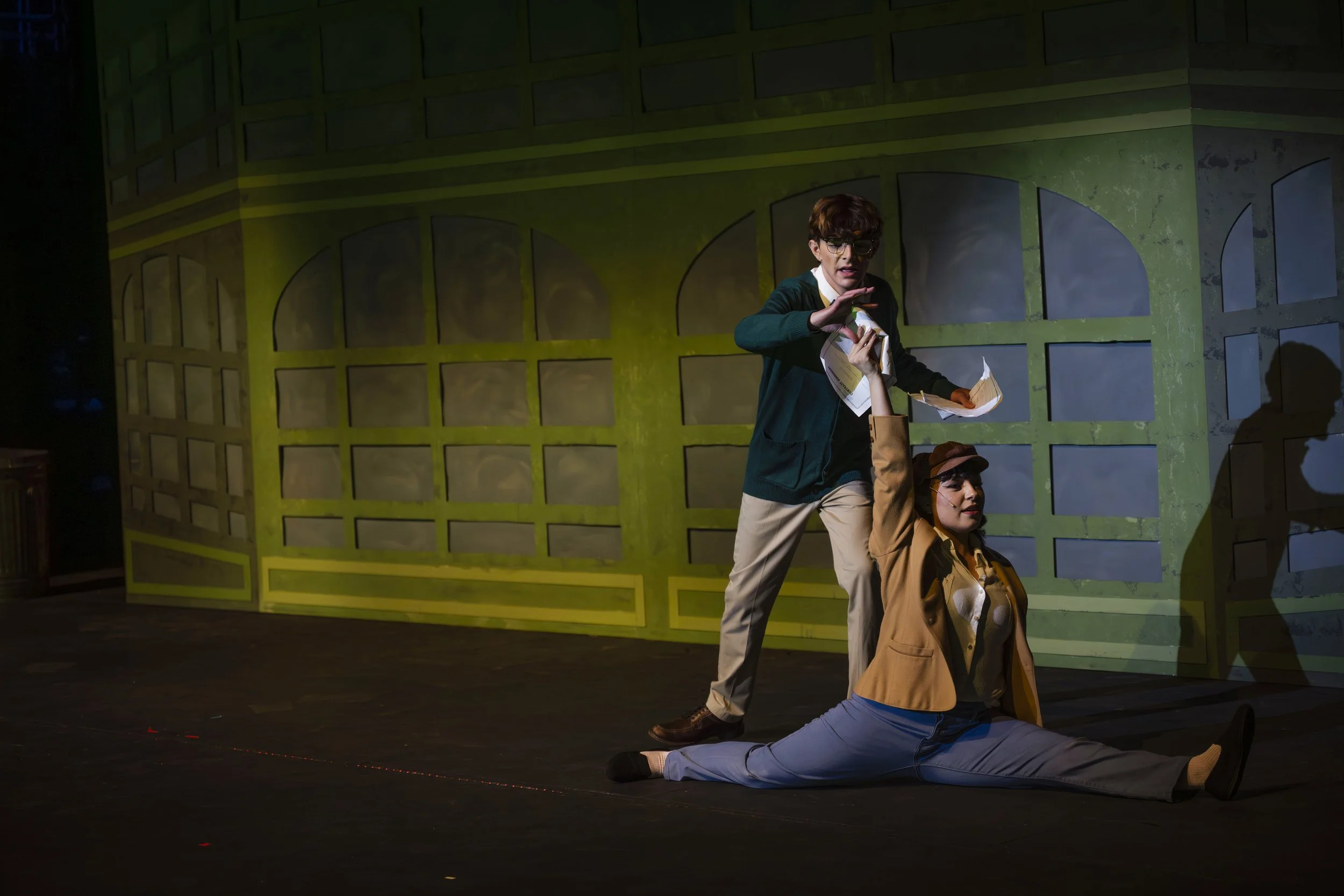 Two actors on stage performing a scene with a green set background, one standing and holding papers and the other seated on the floor, engaging in dramatic expressions.