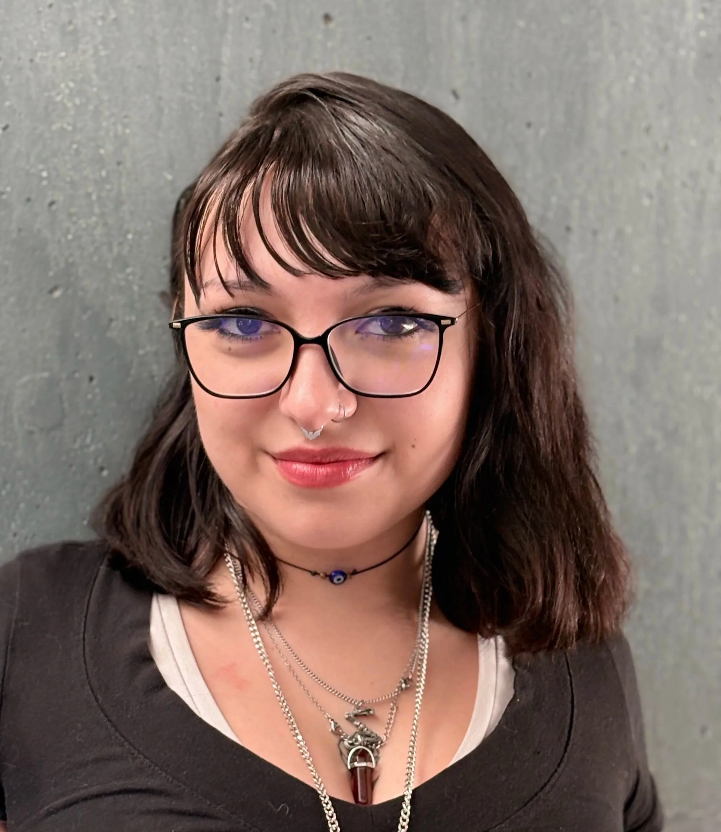 Close-up of a woman with dark brown, wavy hair, wearing black glasses, multiple necklaces, and a nose piercing, looking at the camera against a gray textured background.