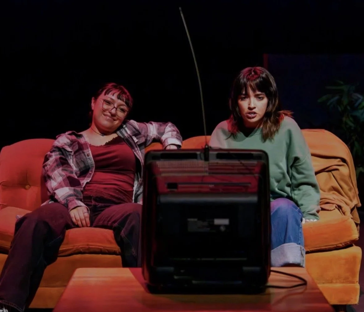 Two young women sitting on an orange sofa, watching television in a dimly lit room, with a vintage radio in front of them.