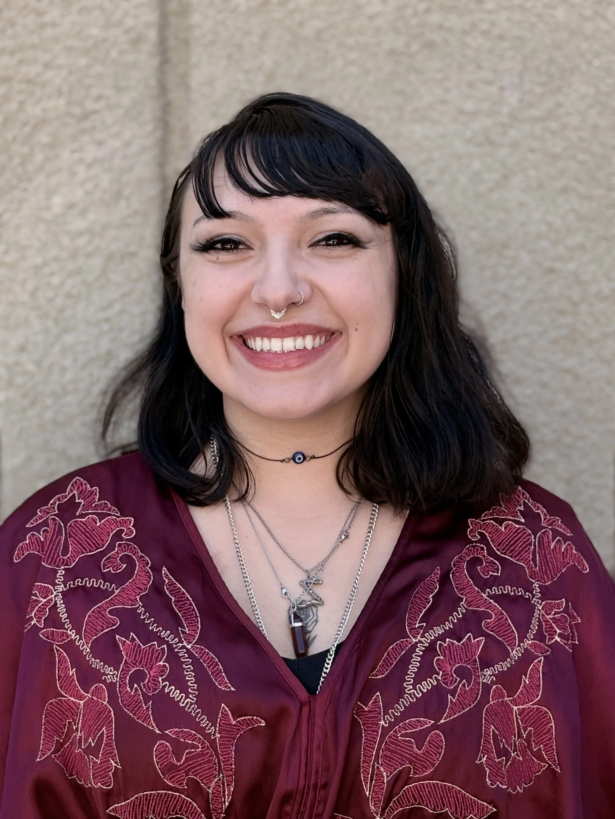 Portrait of a smiling woman with dark hair and bangs, wearing layered necklaces, a nose ring, and a burgundy embroidered top, standing against a beige textured wall.