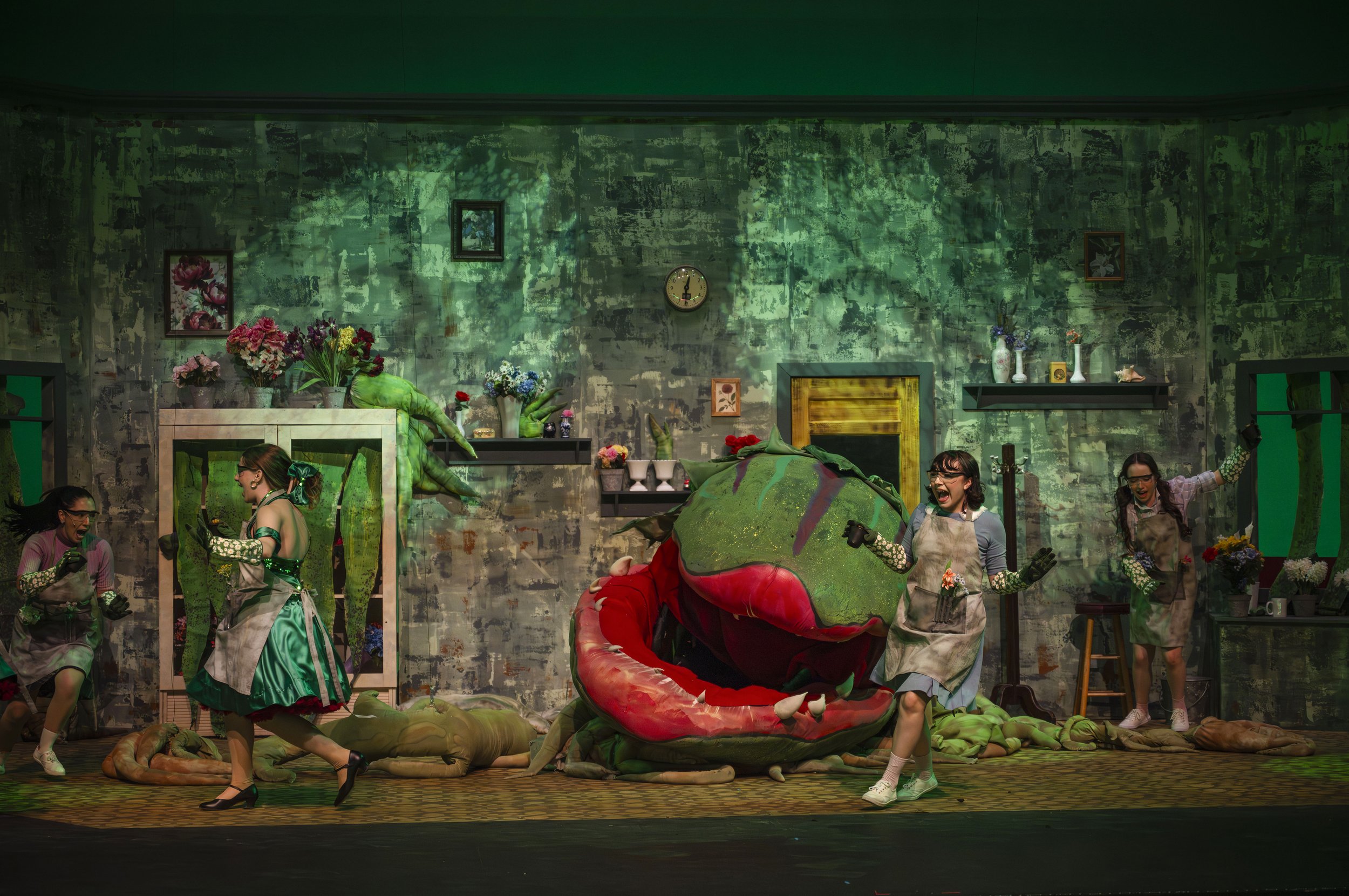 A theatrical stage performance featuring a group of women dressed in green costumes with floral accessories, dancing around a large, colorful dragon prop. The set includes a painted backdrop with framed artwork, plants, and a clock on the wall.