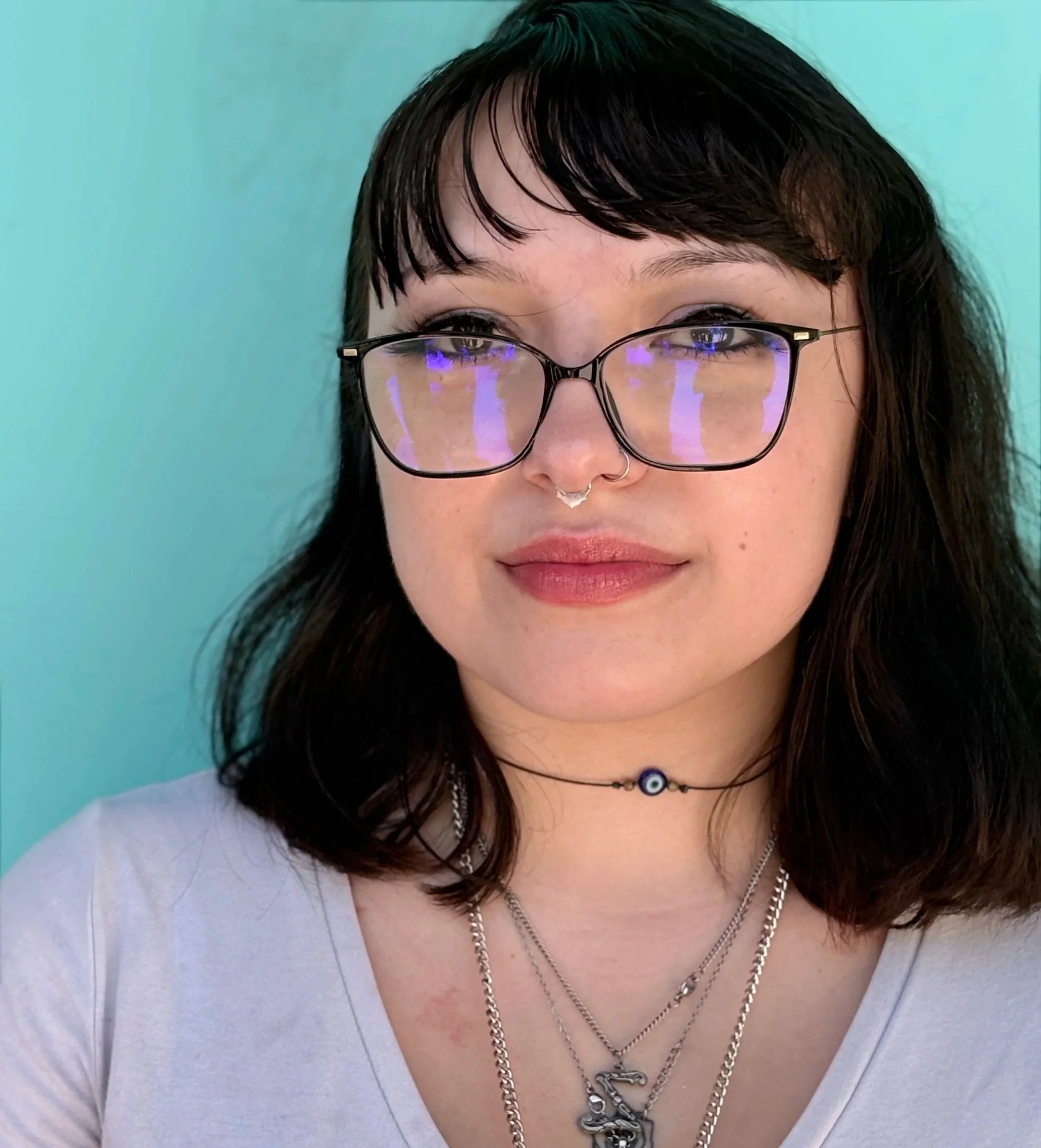 Close-up portrait of a young woman with black hair, wearing glasses with purple reflections, multiple necklaces including a silver chain and an evil eye pendant, and a white shirt against a teal background.
