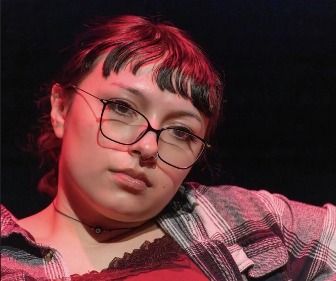 A young person with short, dark hair and glasses, wearing a septum piercing, a choker, and a checkered shirt, looking at the camera against a dark background with red lighting.