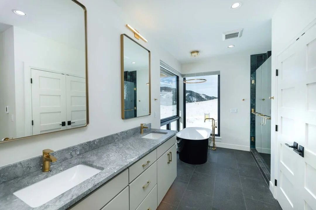 Modern bathroom with dual sinks, large mirrors, a freestanding bathtub near a large window with a view of snowy mountains, and a separate glass shower area.