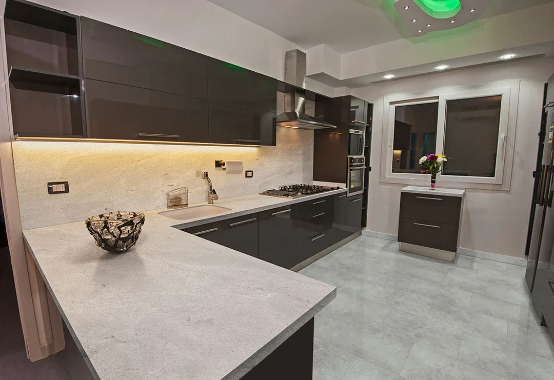 Modern kitchen with gray cabinets, white marble countertops, a gas stove, stainless steel range hood, a double oven, a bowl on the counter, a window with a vase of flowers, and tiled floor.