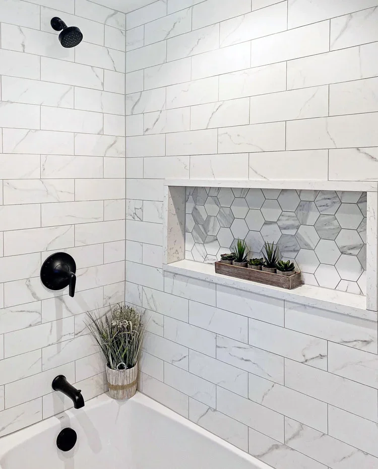 Bathroom shower with white marble tiles, black fixtures, a built-in niche with hexagonal tiles and plants, and a potted plant near the bathtub.