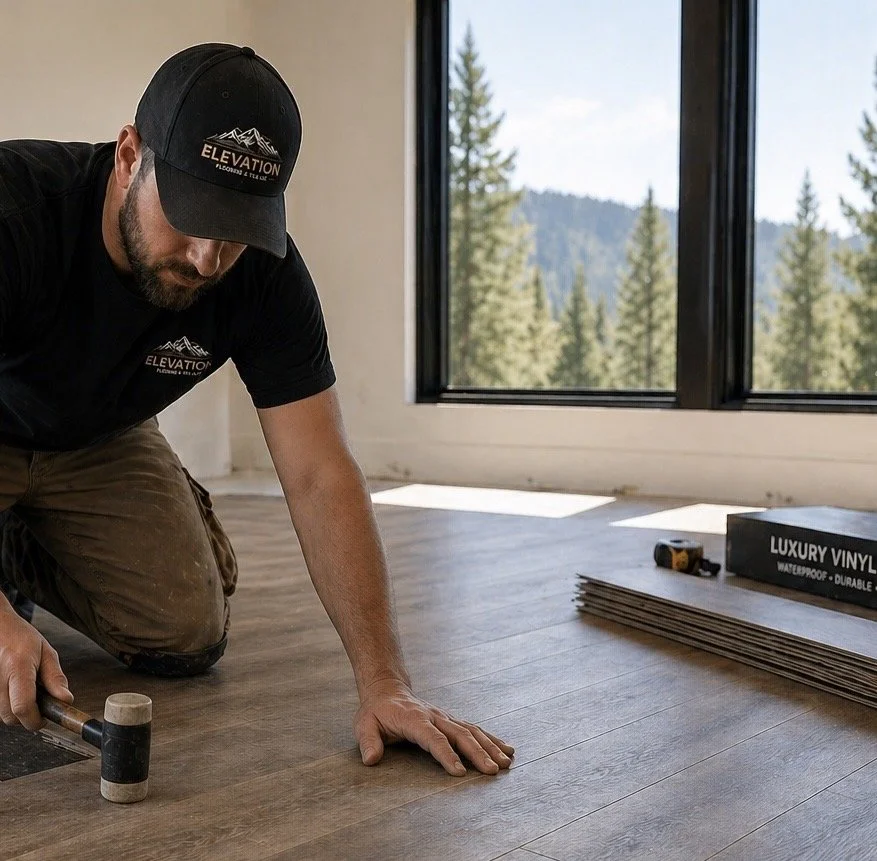 A man installing new wood flooring in a room with large windows showing a forested mountain landscape. He is kneeling, wearing a black cap and t-shirt with 'Elevation' logo, and using a mallet.
