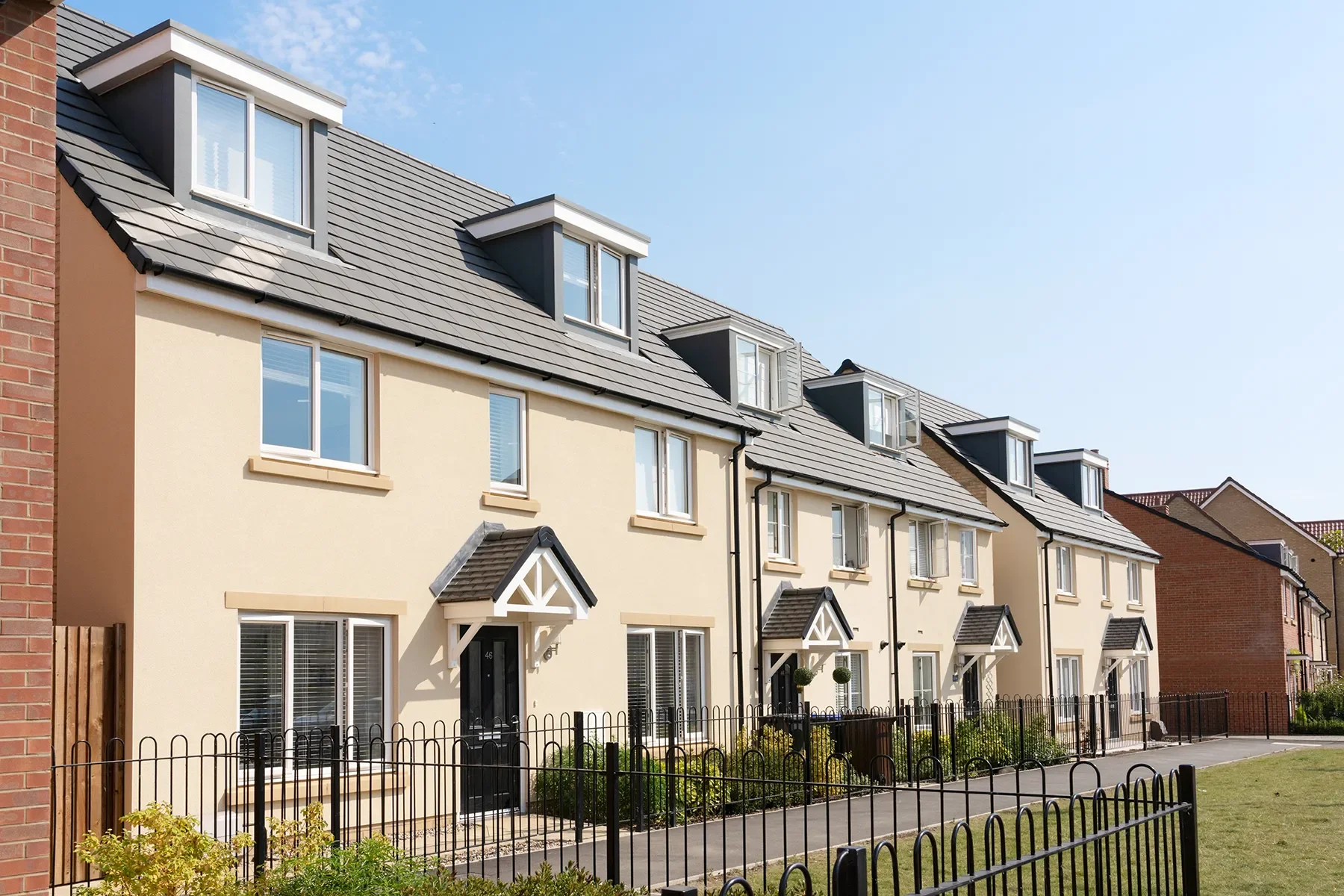 A row of modern, white townhouses with black front doors, black fencing, and multiple windows, with slate roofing and small front yards, under a blue sky.