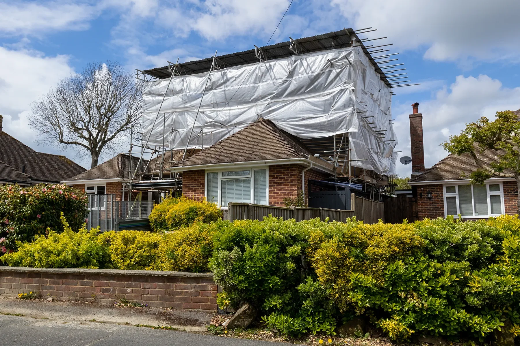 A house under renovation with scaffolding and white protective sheeting on the upper part of the building, surrounded by a garden with green shrubs, a brick wall, and neighboring houses in a suburban neighborhood under a partly cloudy sky.