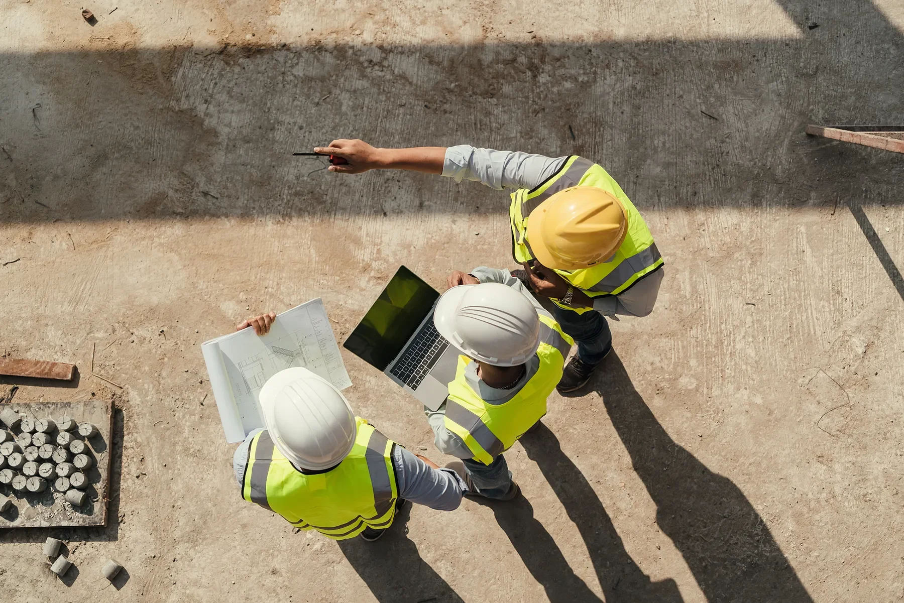 Top-down view of four construction workers wearing yellow and white safety vests and hard hats, reviewing blueprints and a laptop on a construction site with dirt ground and scattered materials.
