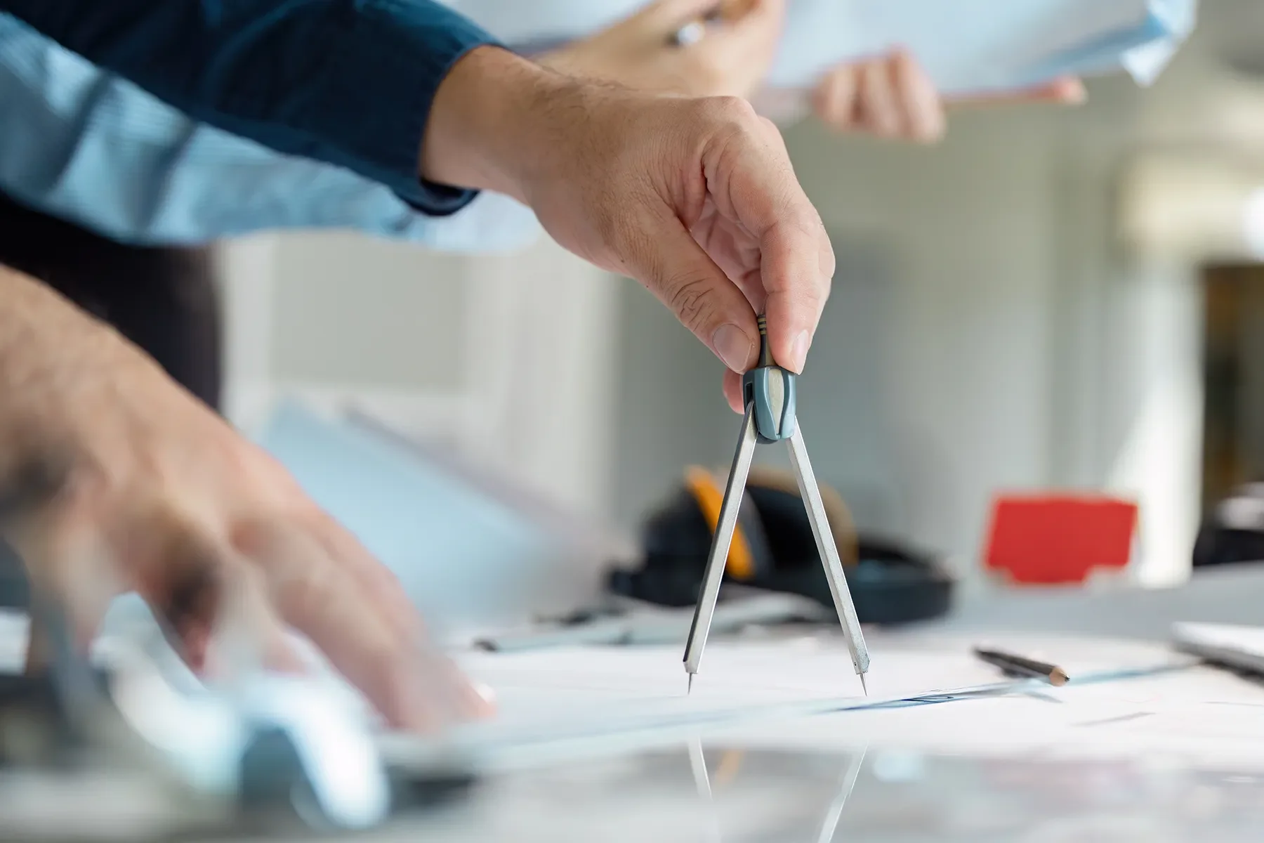 A person uses a brass geometric compass on a piece of paper, with a background of a desk or workspace that includes blurred objects such as a black container, a red item, and writing tools.