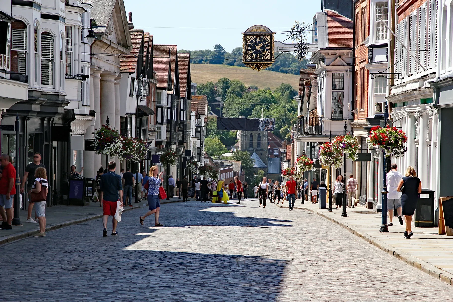 A busy cobblestone street in a historic town lined with shops and buildings decorated with hanging flower baskets, with people walking and shopping on a sunny day.