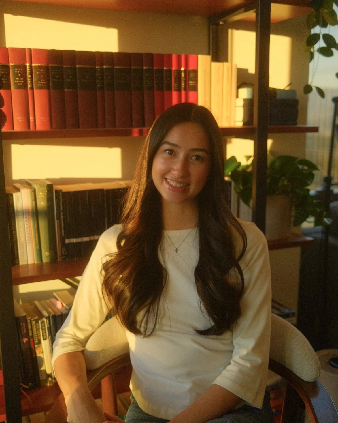 A young woman with long, wavy brown hair, smiling, sitting in front of a bookshelf filled with colorful books, illuminated by warm, golden light.