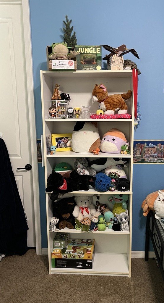 A white bookshelf filled with plush toys and collectibles against a blue wall. The top shelf has a small tree, a plush frog, and some books. The second shelf features stuffed animals, including a large cat, a plush frog, and other toys. The third shelf contains various plush characters, including a large round plush with a pink face, a green turtle, and other small animals. The fourth shelf displays a mix of plush toys and small figures, including a panda, elephant, and other animal plushies. The bottom shelf holds a Monopoly game box, and there are more plush toys on the floor.