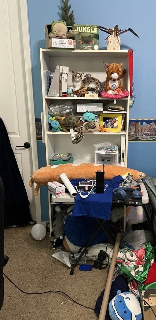 A cluttered room with a white bookshelf filled with stuffed animals and decorative items. There's a desk covered with various objects, a blue cloth, a pillow, and an office chair. The background shows a blue and gold wall, and the floor has miscellaneous items including a balloon and a rolled-up poster.