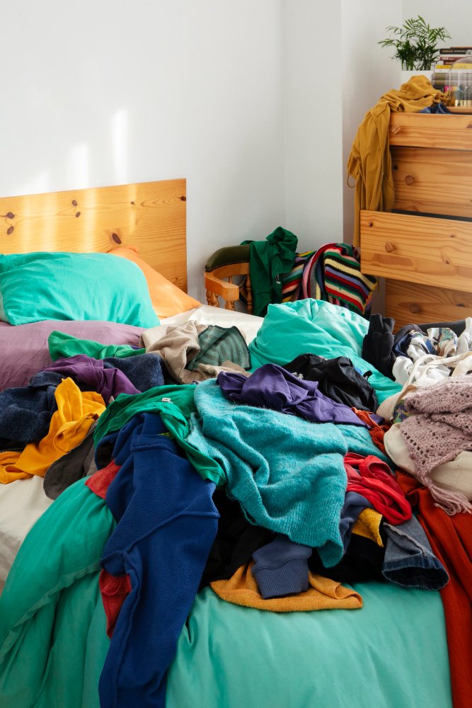 Messy bed with colorful clothes and blankets, wooden headboard, dresser with books and a plant in the background.