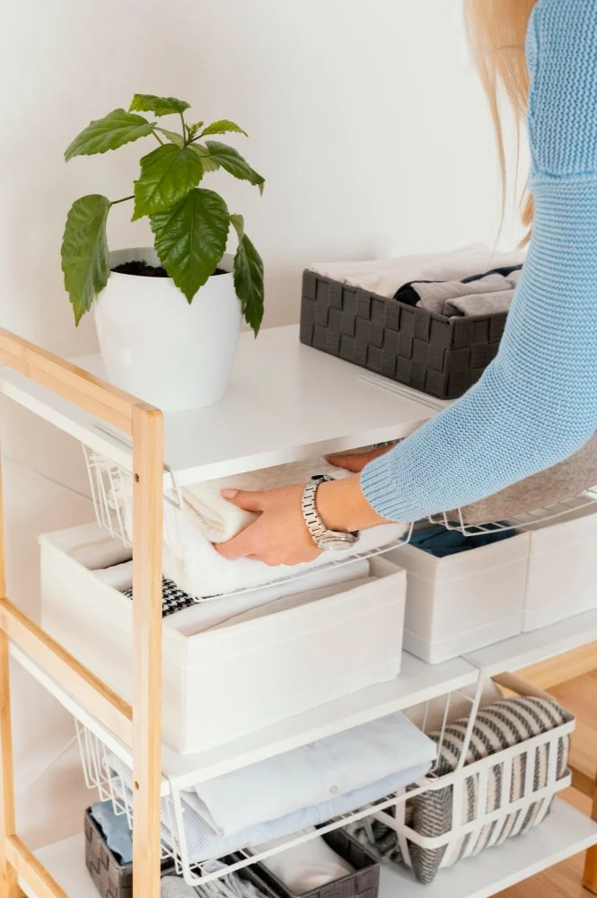 Person folding a white towel on a white shelf unit with neatly organized baskets and a potted green plant on top.