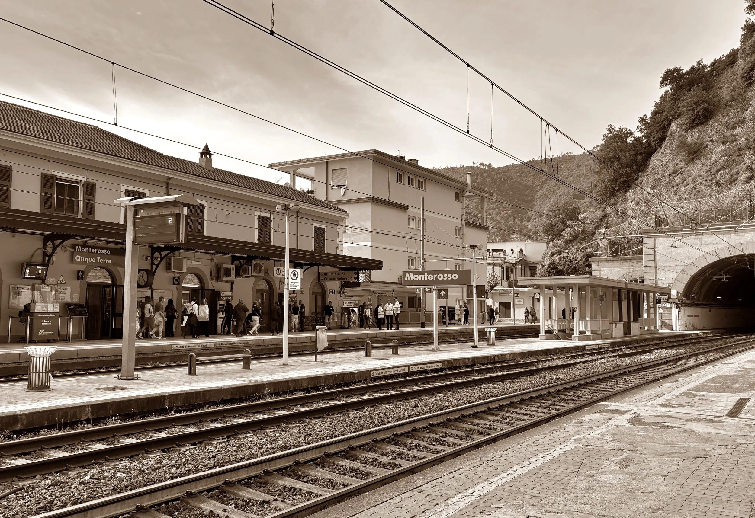A train station platform with tracks in the foreground, a building with shops and a sign that says Monterosso in the background, and people waiting for a train. Overhead wires are visible above the platform, and there are hills covered with trees behind the buildings.