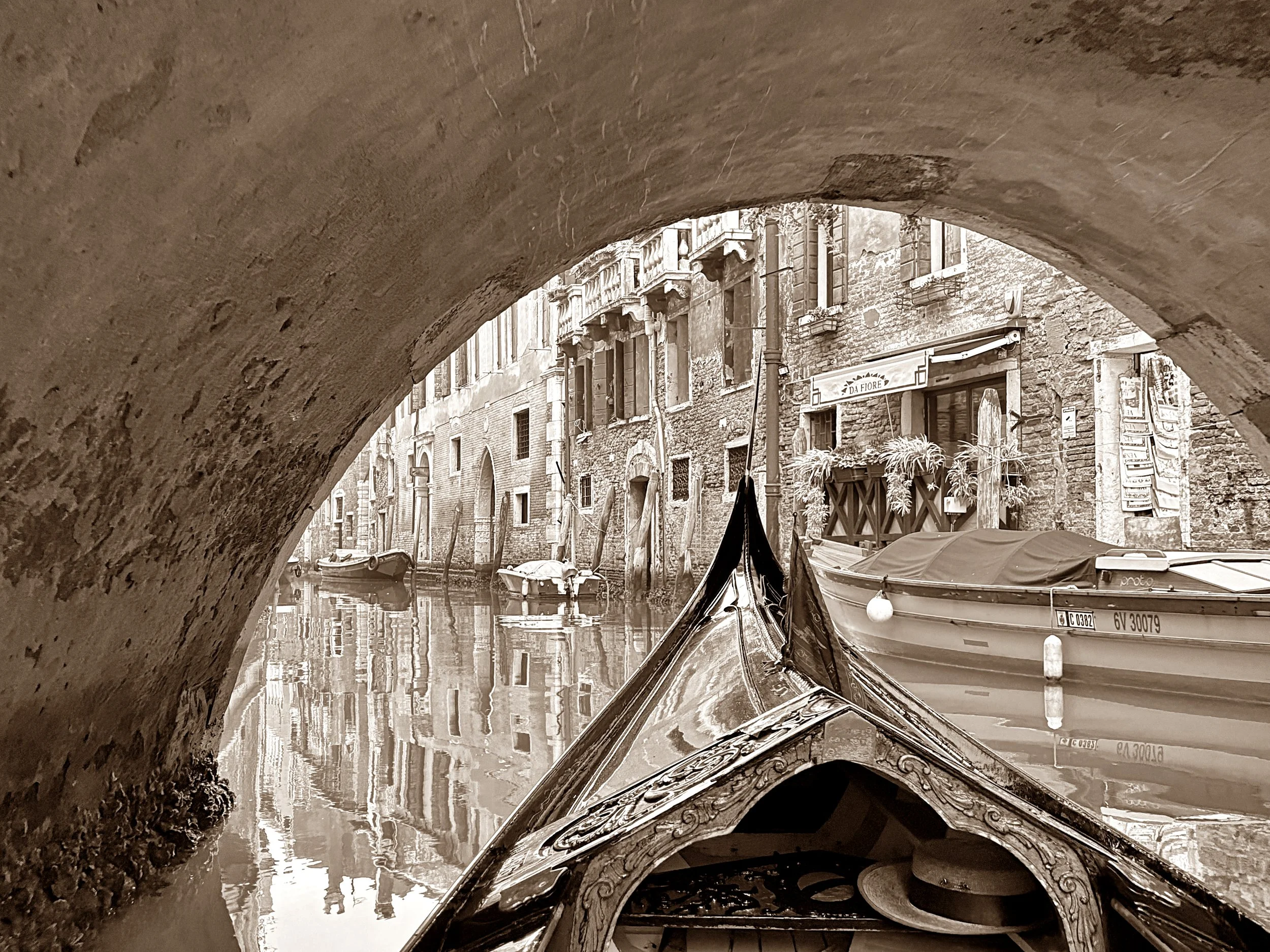 A canal scene in Venice, Italy, viewed through a stone arch bridge. Several boats are docked along the narrow waterway, with old brick buildings featuring balconies and shuttered windows on either side.