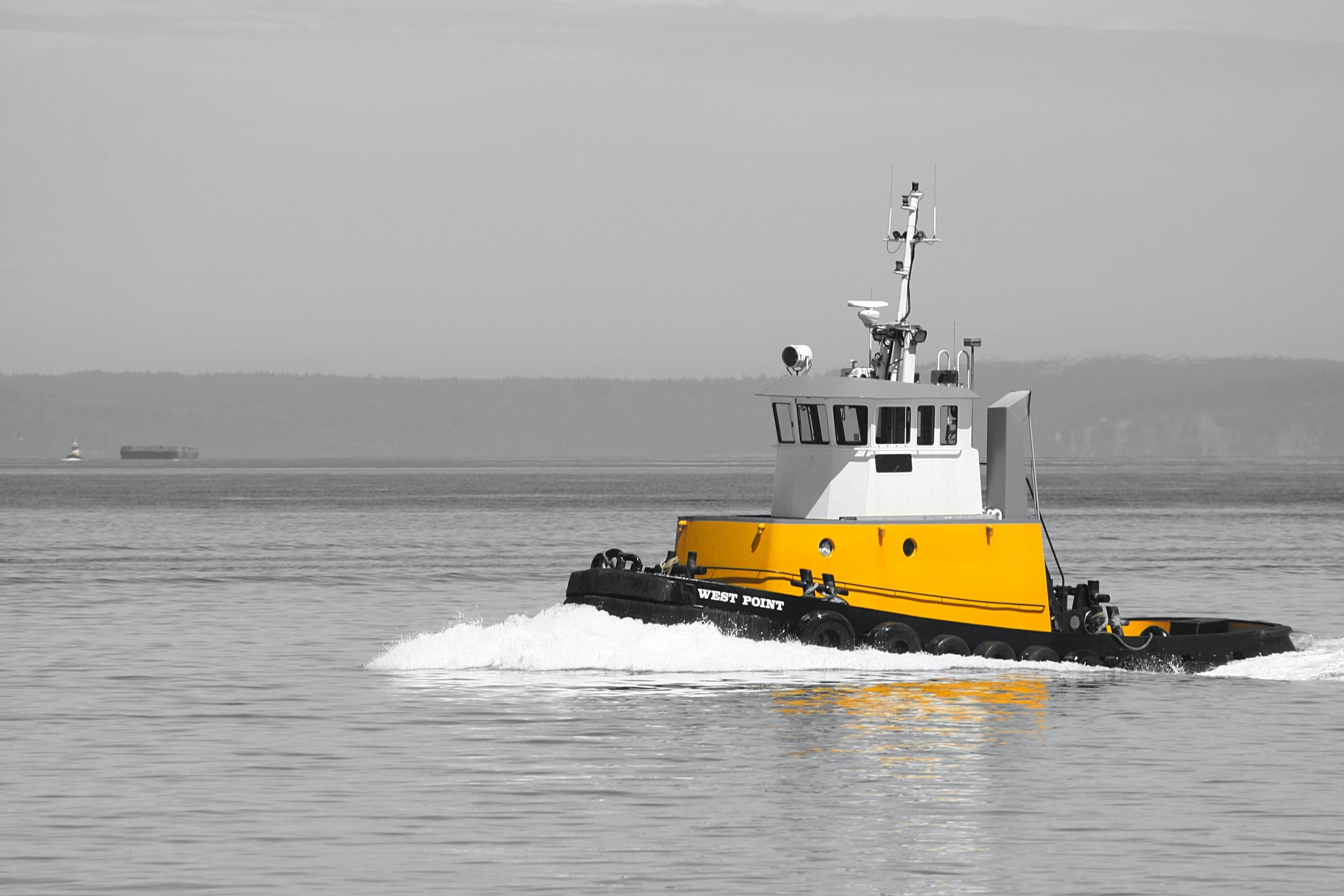 A yellow tugboat named West Point moving through water with a distant shoreline and ships in the background during daytime.