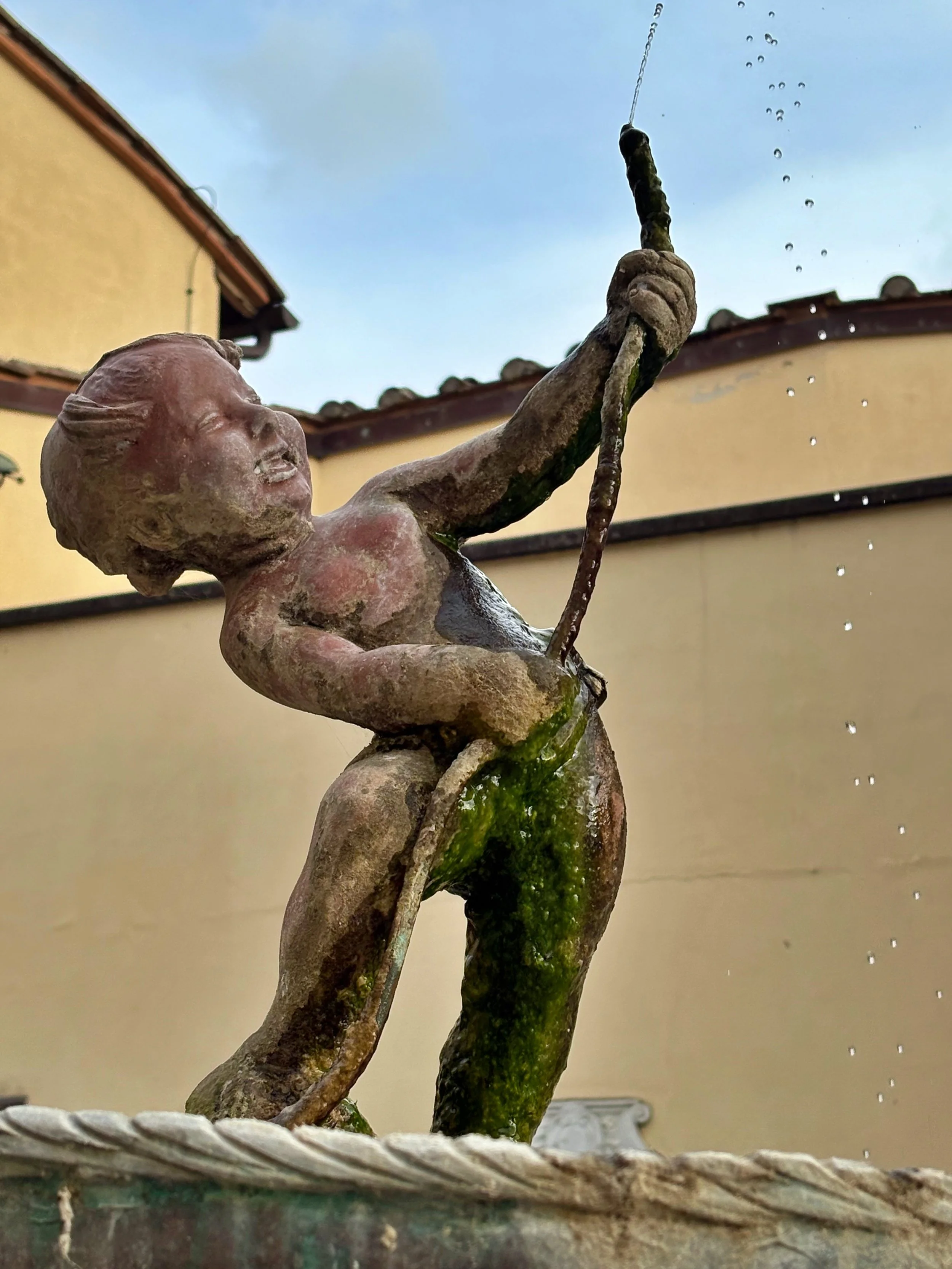 A weathered sculpture of a child with a smiling facial expression, holding a fishing rod with a line going into the sky. The sculpture appears aged with moss on the surface and is placed on a woven basket-like base, with a residential building and a cloudy sky in the background.